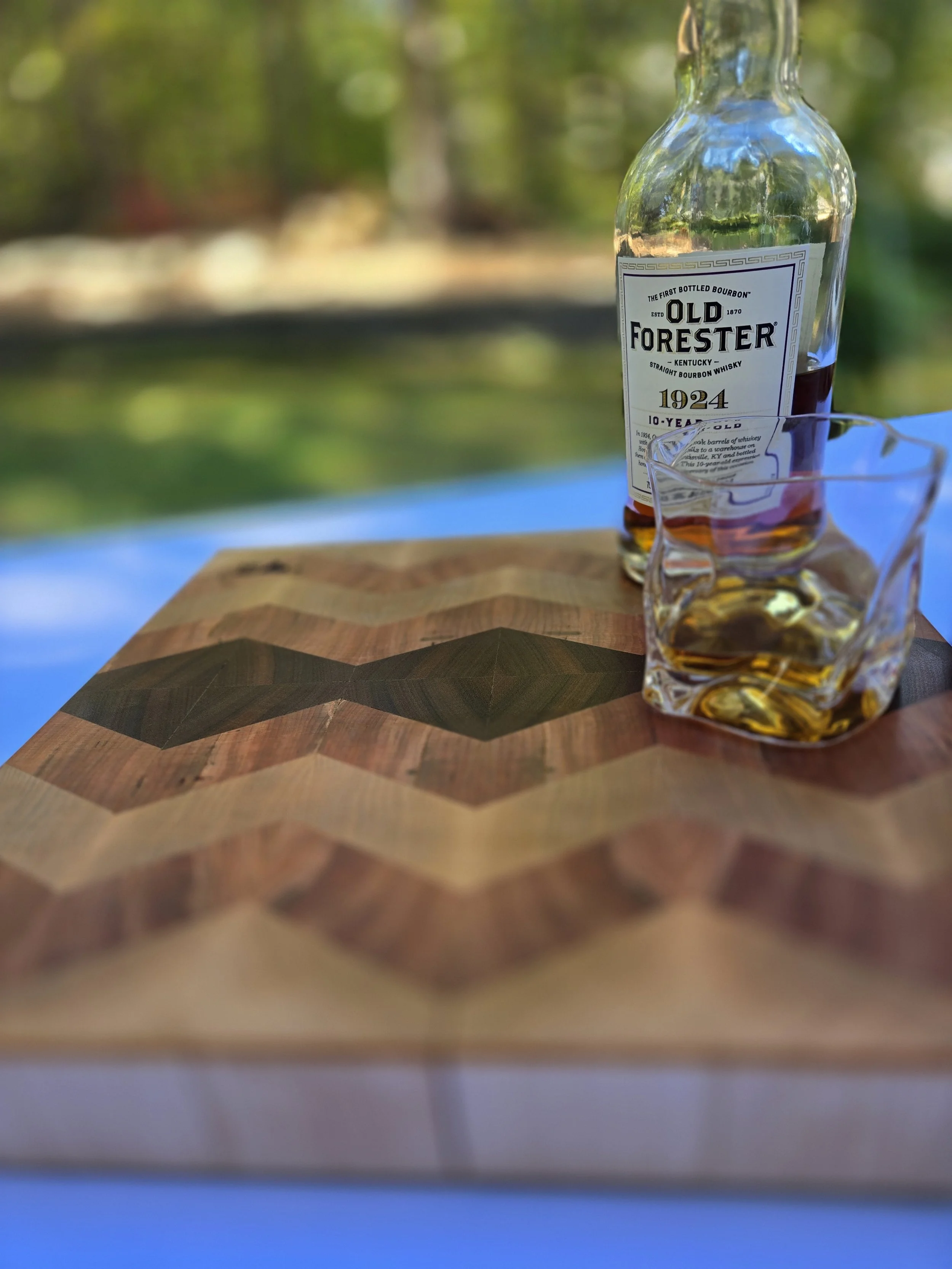 A wooden table with a patterned inlay, a bottle of Old Forester bourbon, and a glass with bourbon and ice cubes outdoors, with a blurred background of trees and greenery.