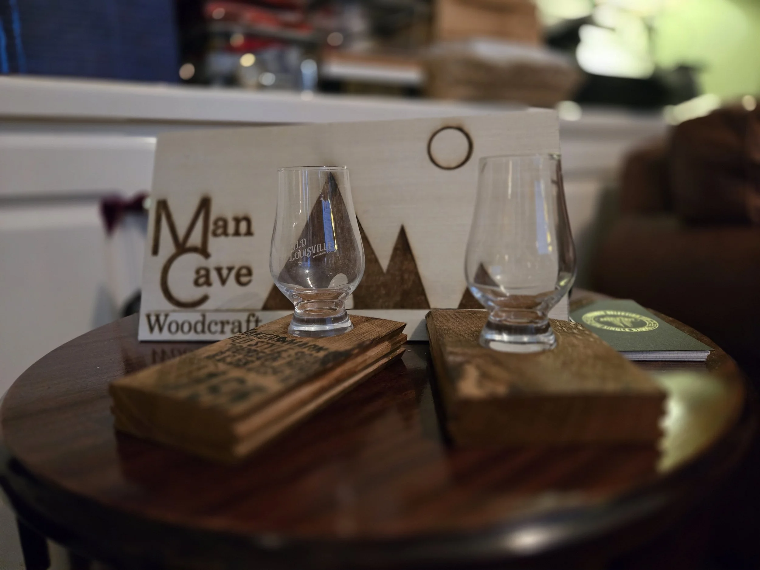 Two empty whiskey glasses placed on small wooden coasters on a round wooden table. Behind the glasses, a sign with the words 'Man CaveWoodcraft' is visible, and there are stacks of business cards or pamphlets to the right. The background is blurred w