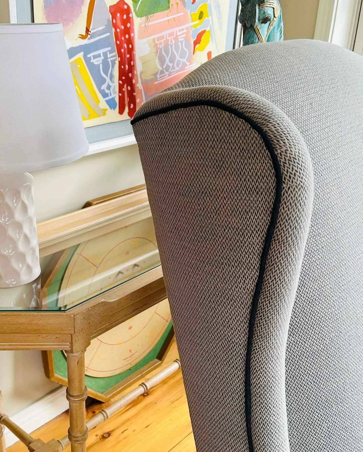 The image shows a part of a cozy living room corner. There is a gray upholstered armchair with black piping, a wooden and glass side table with a framed artwork featuring a colorful, abstract painting of a bird on the wall behind. A white textured lamp with a rounded base is placed on the side table.