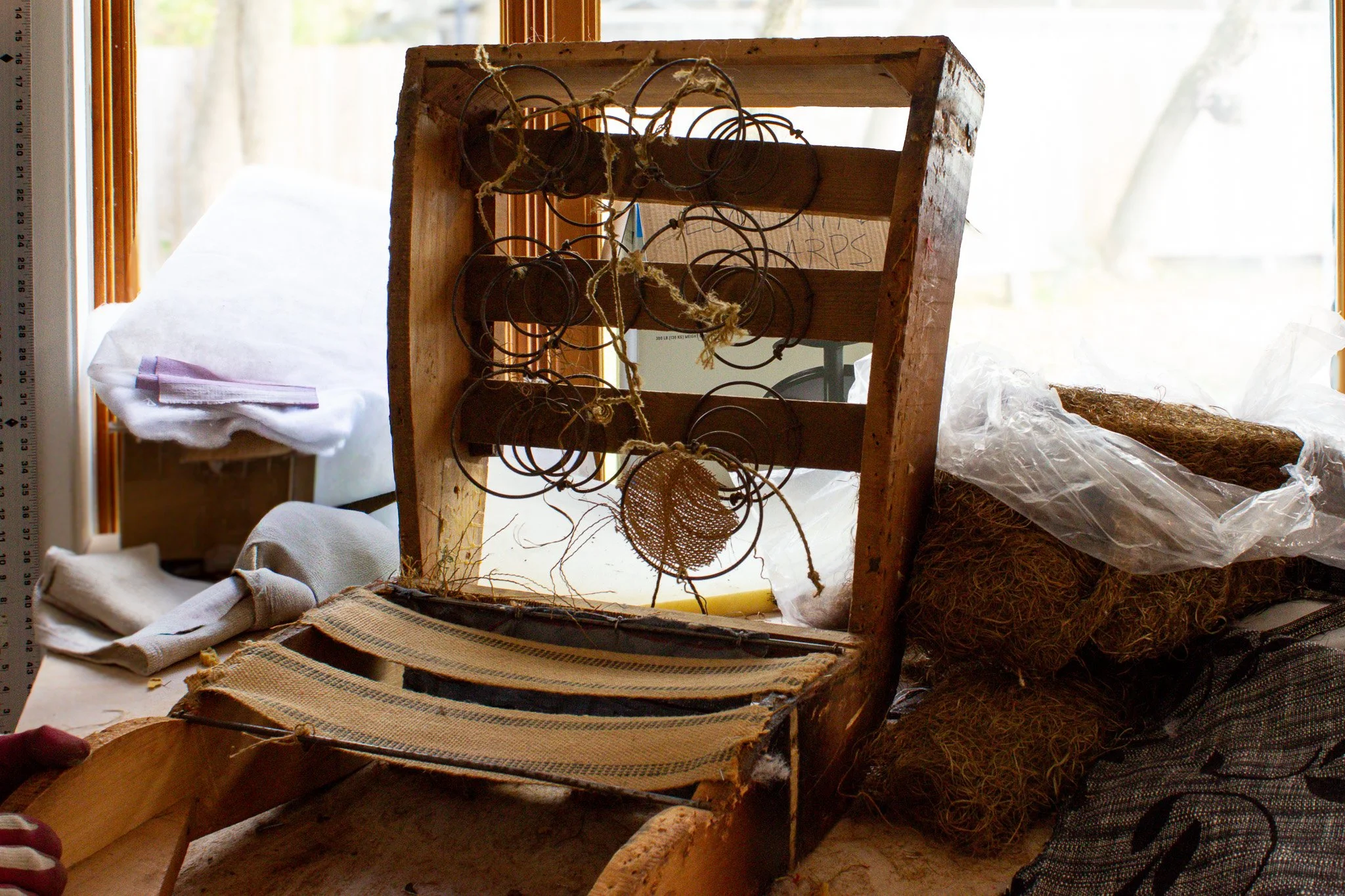 Old wooden loom with metal cycles, some wrapped in string, placed on a table in front of a window. Nearby are clay balls wrapped in plastic, a pair of white gloves, and cloth.