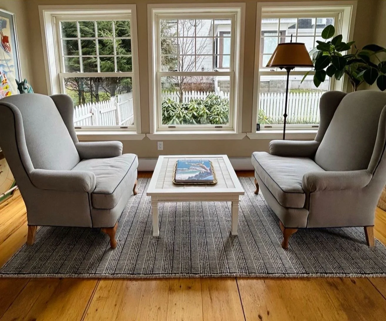 Sunlit living room with two beige armchairs facing each other, a white coffee table between them with a tray holding magazines, and large windows showing a fenced yard with trees and bushes outside.