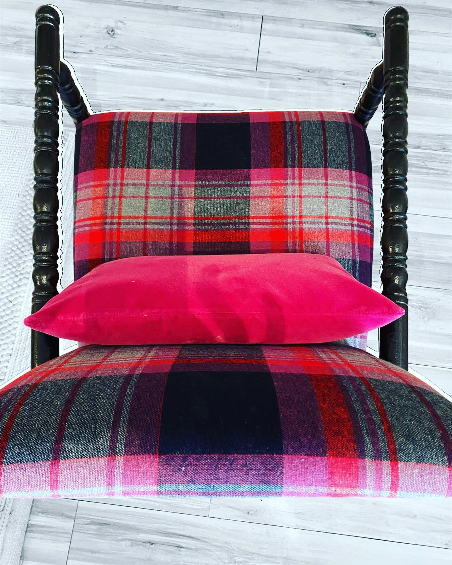 Top-down view of a vintage chair with a red, black, and green plaid fabric and a bright pink pillow on it, against a light wood floor background.