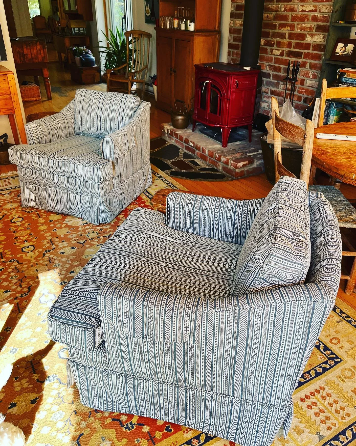 Living room with two striped armchairs, a patterned rug, a red wood stove, and brick fireplace. There's a dining table with chairs and a bookshelf.