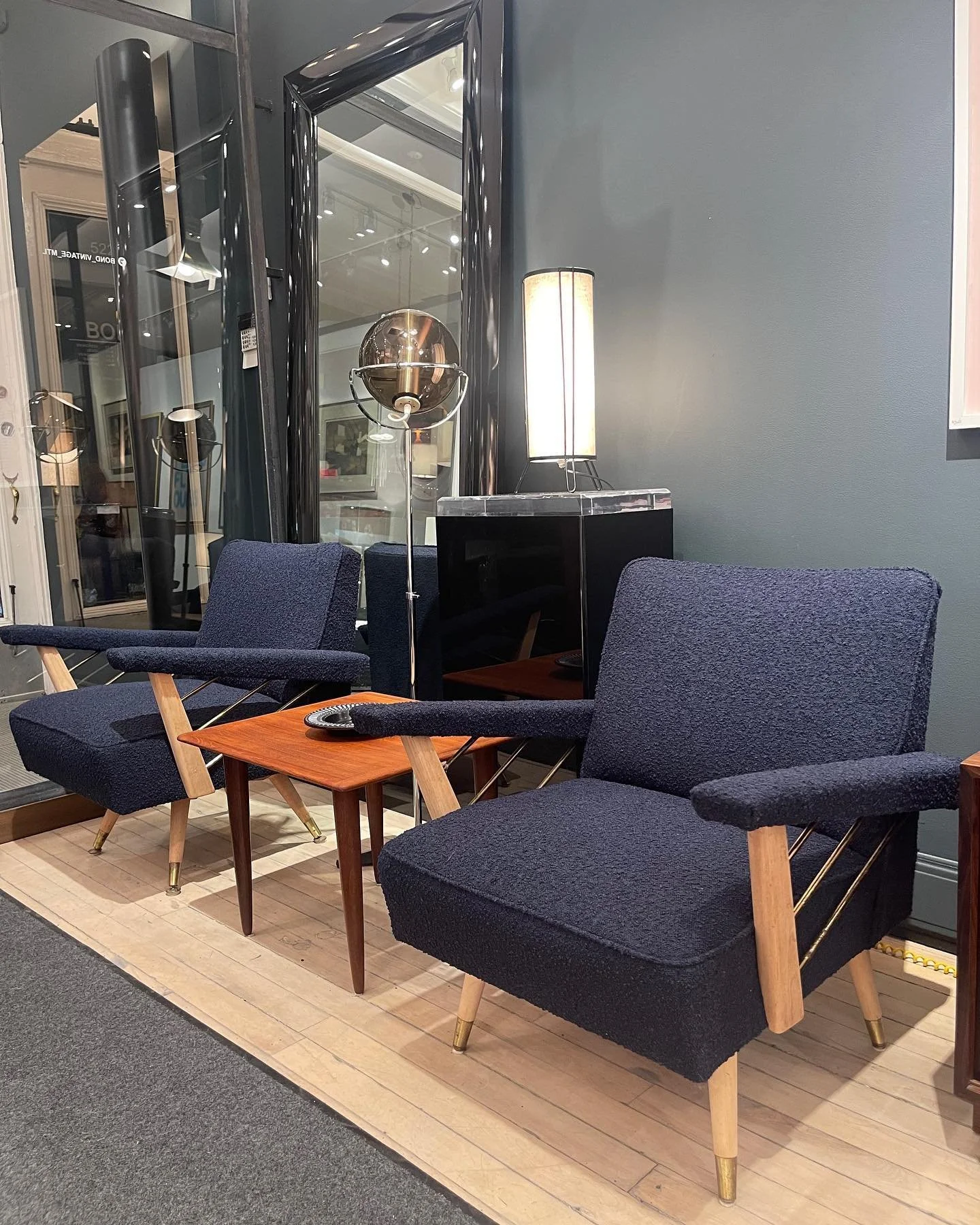 Mid-century modern lounge area with two navy upholstered armchairs with wooden and brass legs, a small wooden side table with a black ashtray on top, a tall white floor lamp, and a black reflective cabinet with a marble top against a gray wall. A lar