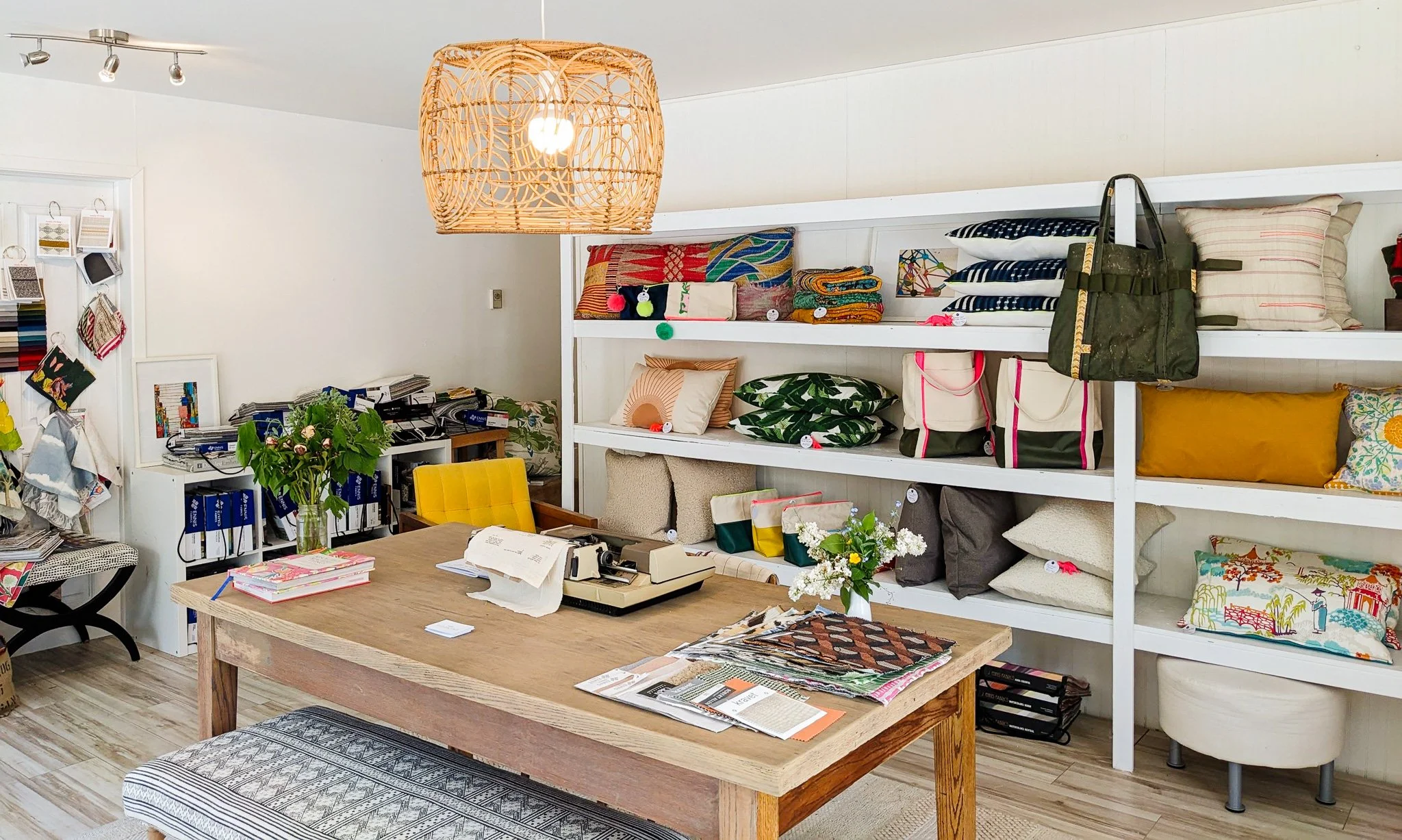Interior of a home decor store with a wooden table, a yellow chair, shelves filled with pillows, tote bags, and textiles, decorative flowers, and a woven pendant light