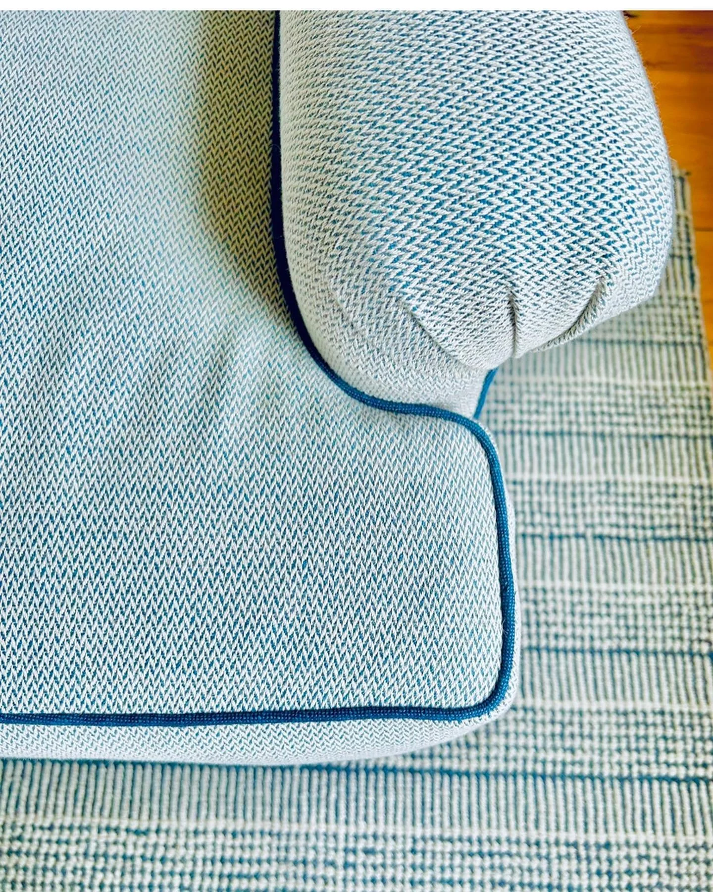 Close-up of a patterned upholstered chair with a rounded armrest and a blue piping edge, situated on a striped rug.