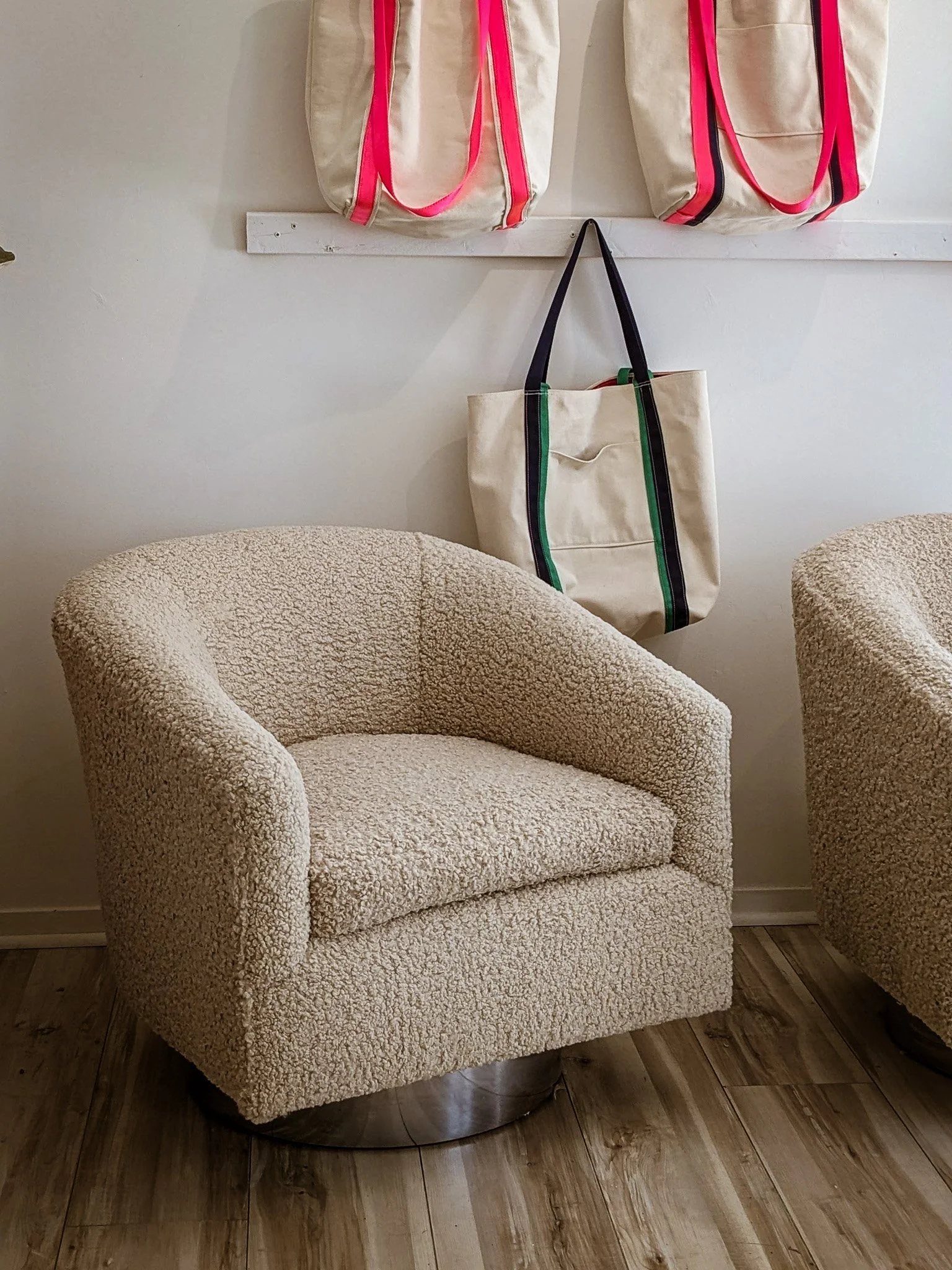 A beige, textured armchair with a rounded back and armrests in a cozy interior, with two more similar chairs partially visible on either side, against a white wall. Two large canvas tote bags with pink and black accents are hanging from a wall-mounte