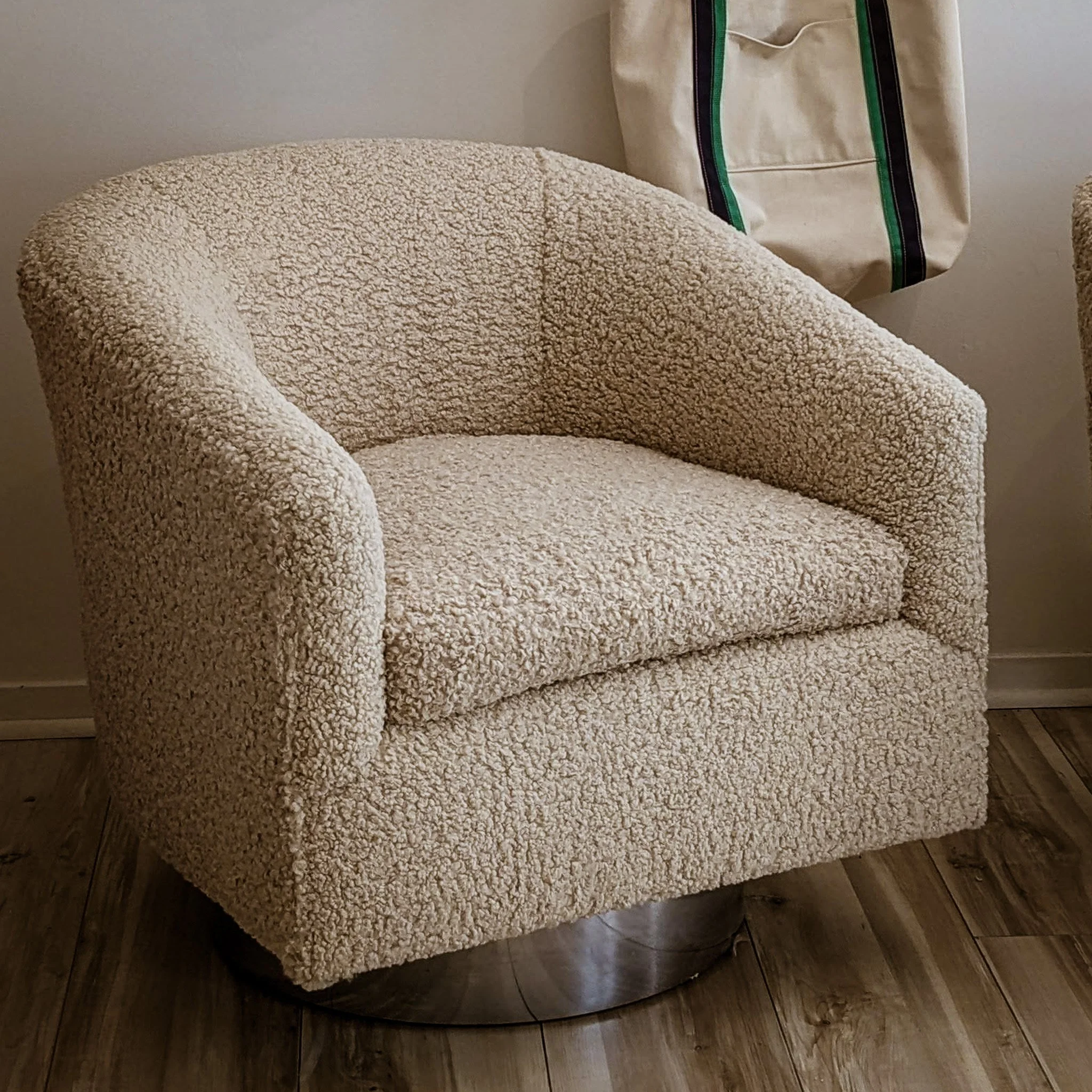 Beige textured upholstered armchair with circular wooden base, placed on wood flooring and partially visible beige tapestry in background.