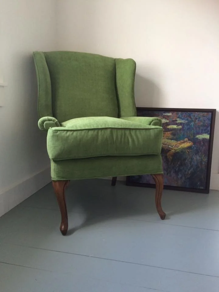 A green upholstered wingback armchair with wooden legs is sitting on a light-colored floor, leaning against a white wall. A framed painting of water lilies and a pond is resting on the floor beside the chair.