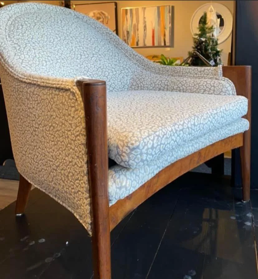 A vintage mid-century modern loveseat with wooden legs and arms, upholstered in a beige and white animal print fabric, positioned on a dark wood floor in a decorated room with artwork and a small Christmas tree in the background.