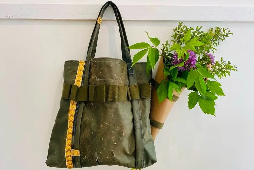 A weathered olive green tote bag with black handles, decorated with a yellow patterned strap, hanging on a white wall. Next to it is a bouquet of purple and green flowers wrapped in brown paper.