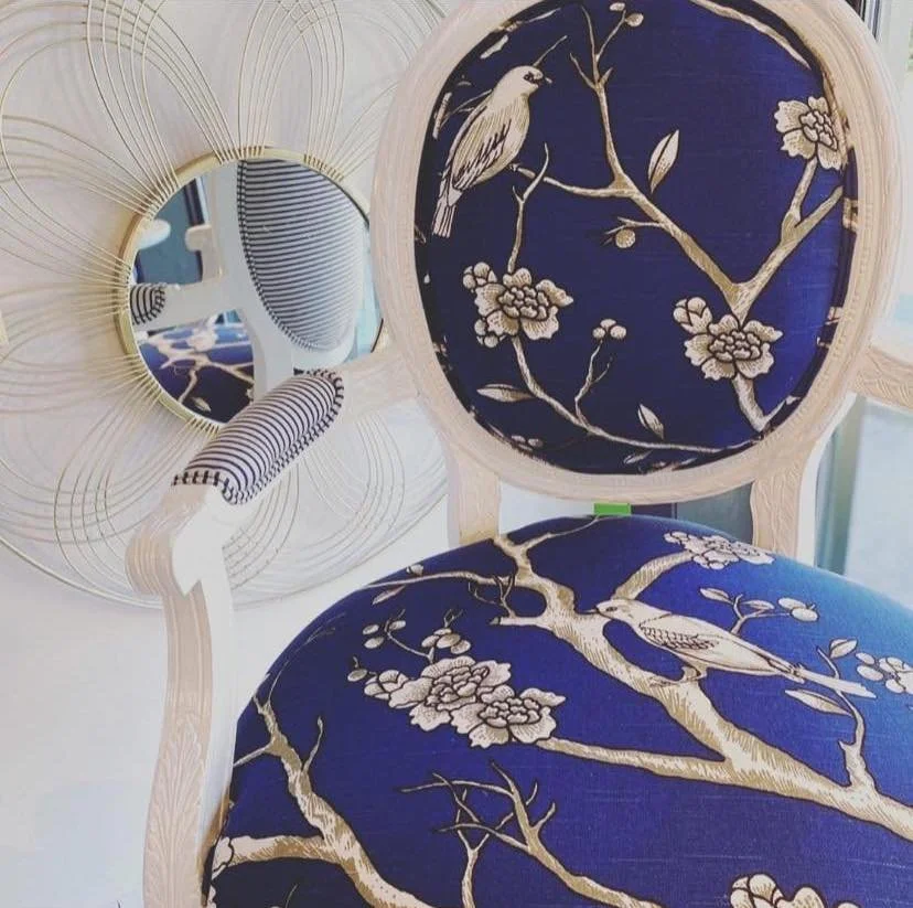A chair with a blue cushion and backrest featuring a floral and bird pattern, placed in front of a round mirror with a decorative frame.