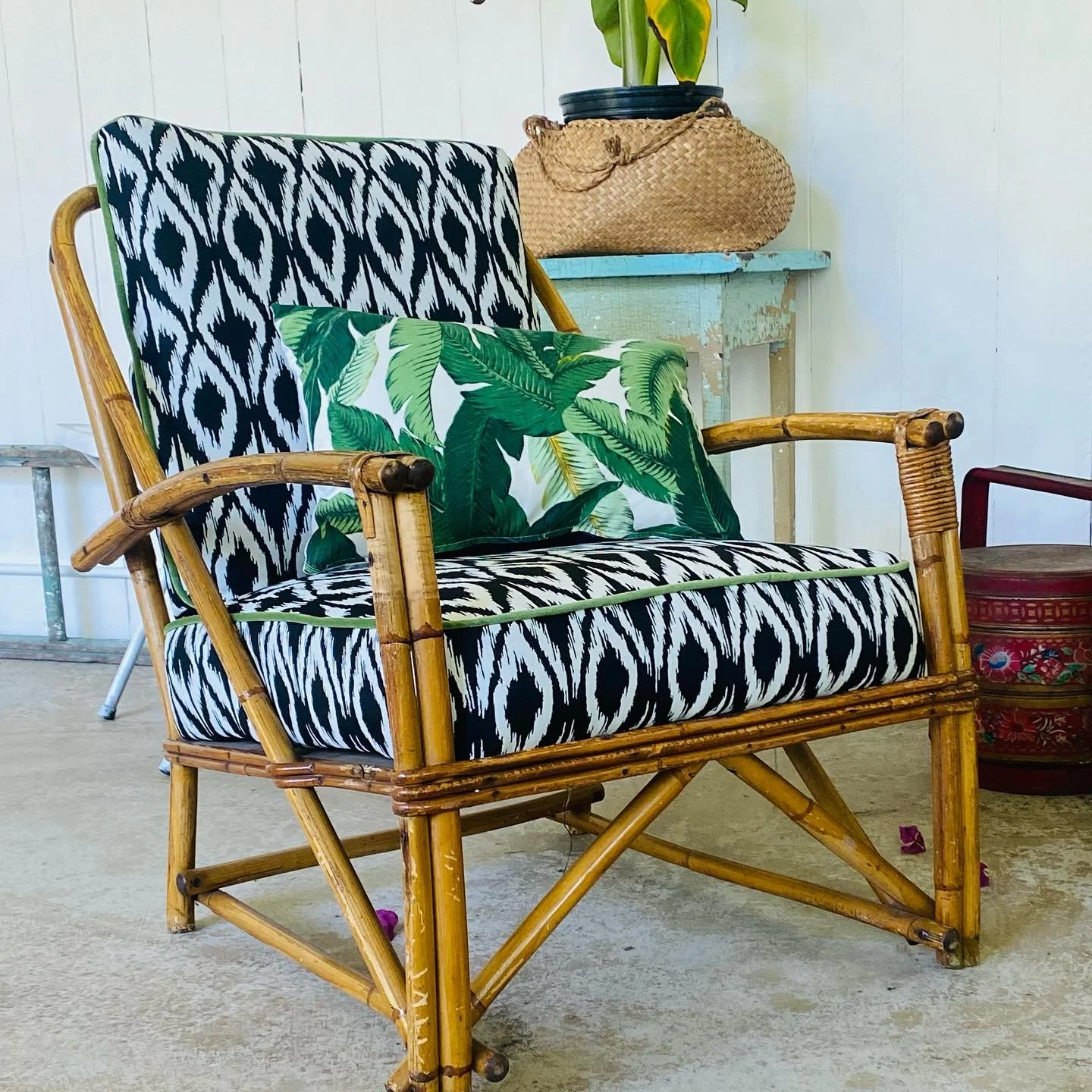 Rattan armchair with black and white patterned cushions and a tropical leaf cushion, positioned in a room with a distressed light blue table in the background, a potted plant, and a decorative basket.