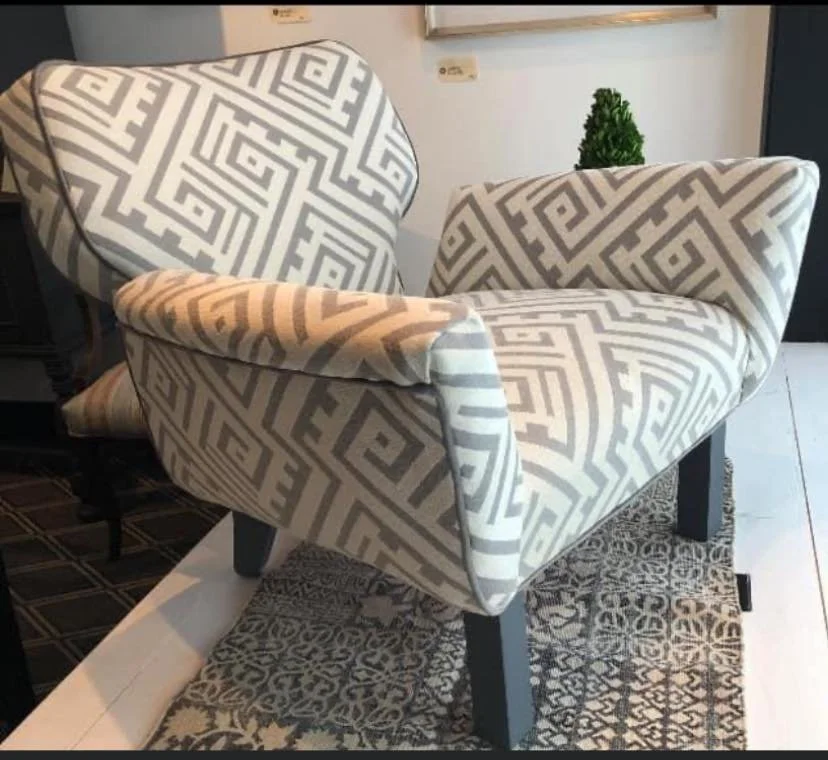 A patterned armchair with beige and gray geometric design, placed on a small rug, with a side table and wall decor in the background.