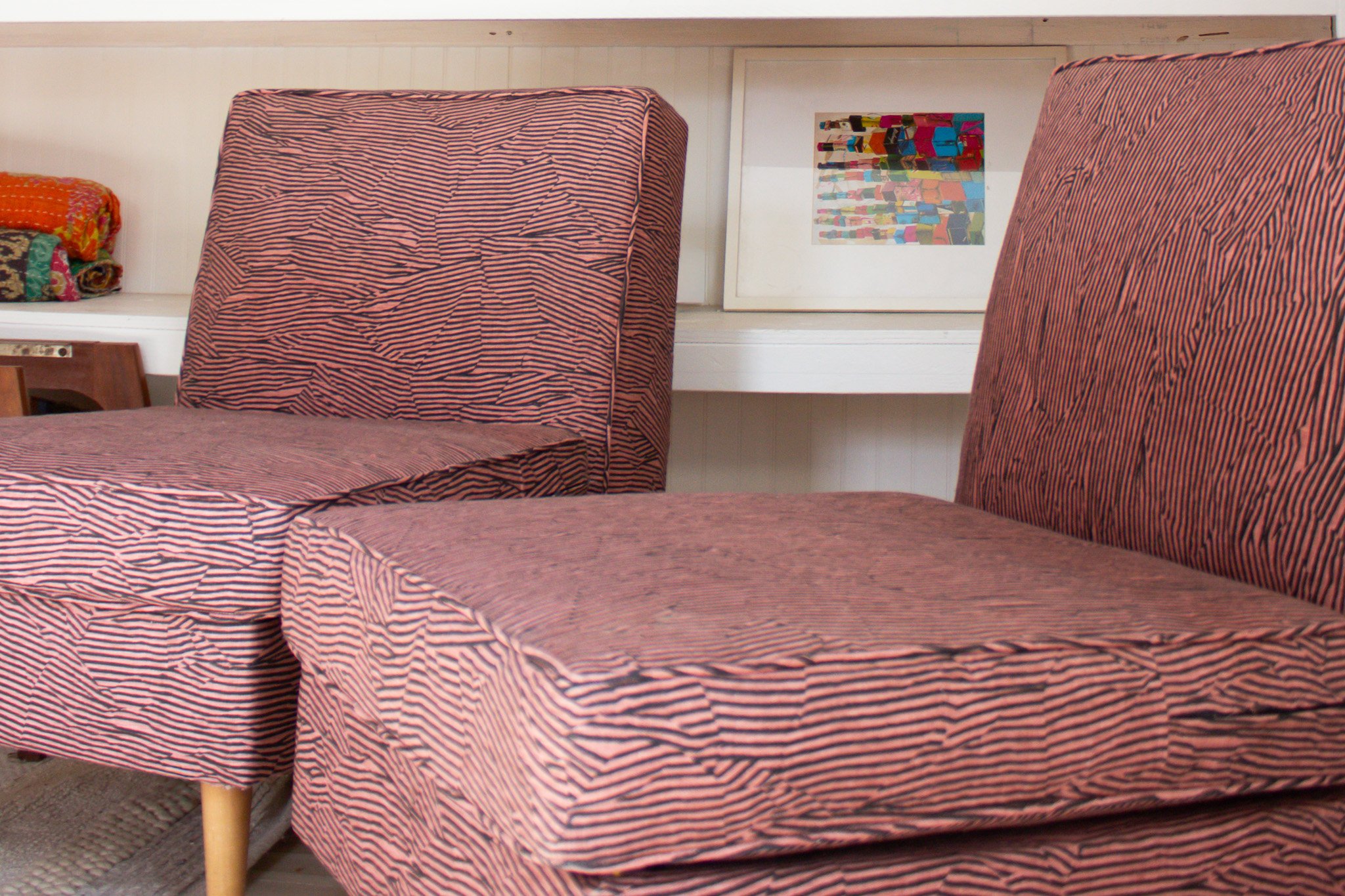 Two chairs with red and black striped fabric and wooden legs in a room with white walls; a framed abstract artwork on the wall behind them.