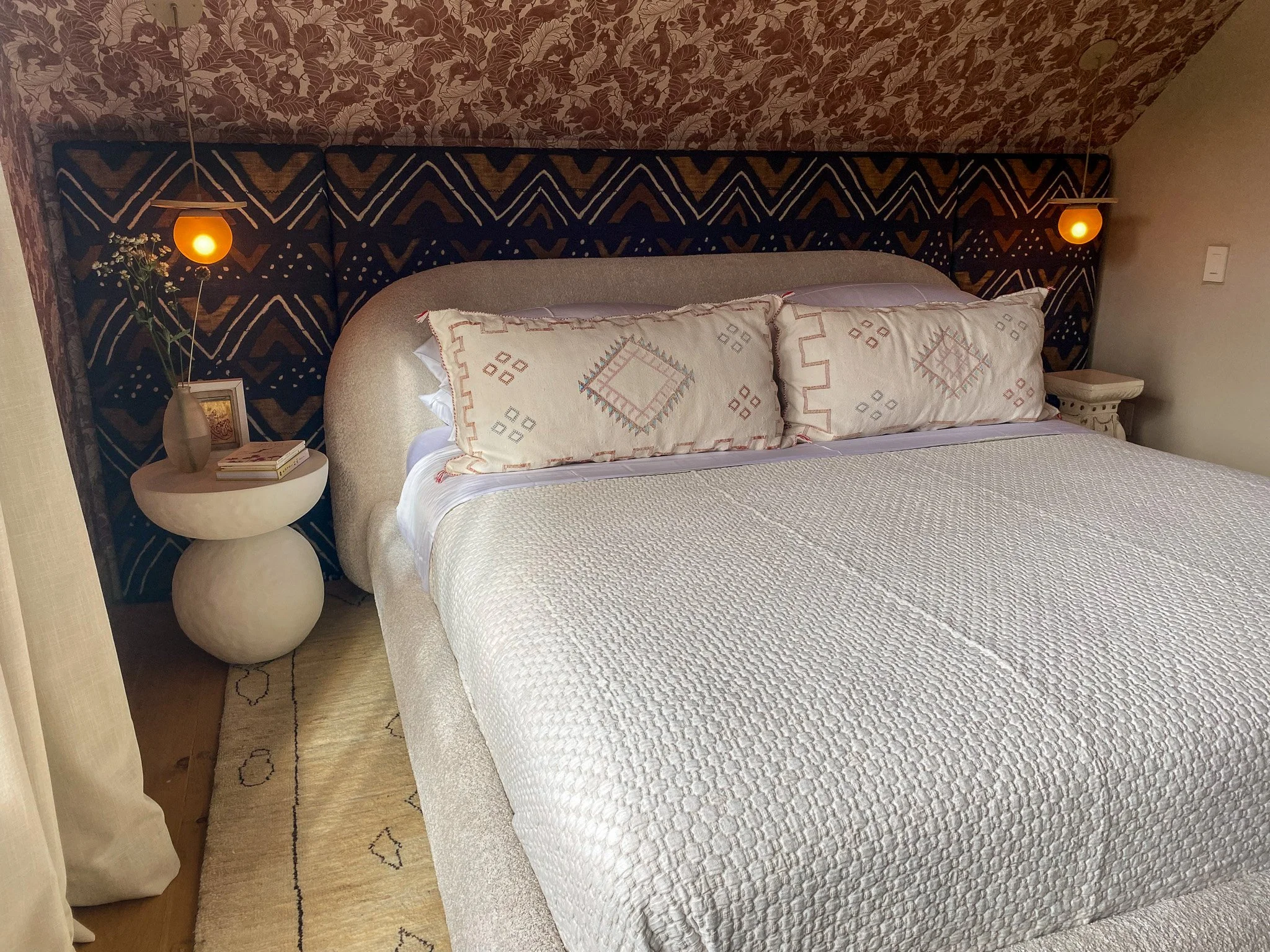 A cozy bedroom featuring a bed with white bedding and embroidered pillows. There is a nightstand with a vase and books, and wall-mounted orange lamps on either side. The headboard has a colorful geometric pattern and the room has warm lighting.