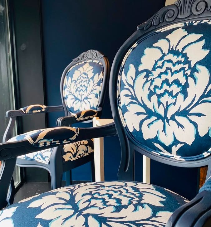 Three wooden chairs with intricate carved designs and blue and white floral upholstery, arranged in a row against a blue wall.