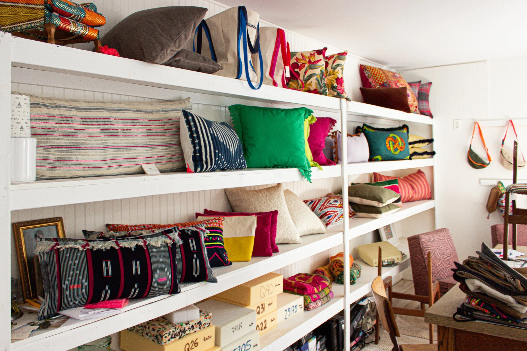 Shelves filled with colorful pillows and bags in a room, with chairs and a table nearby.