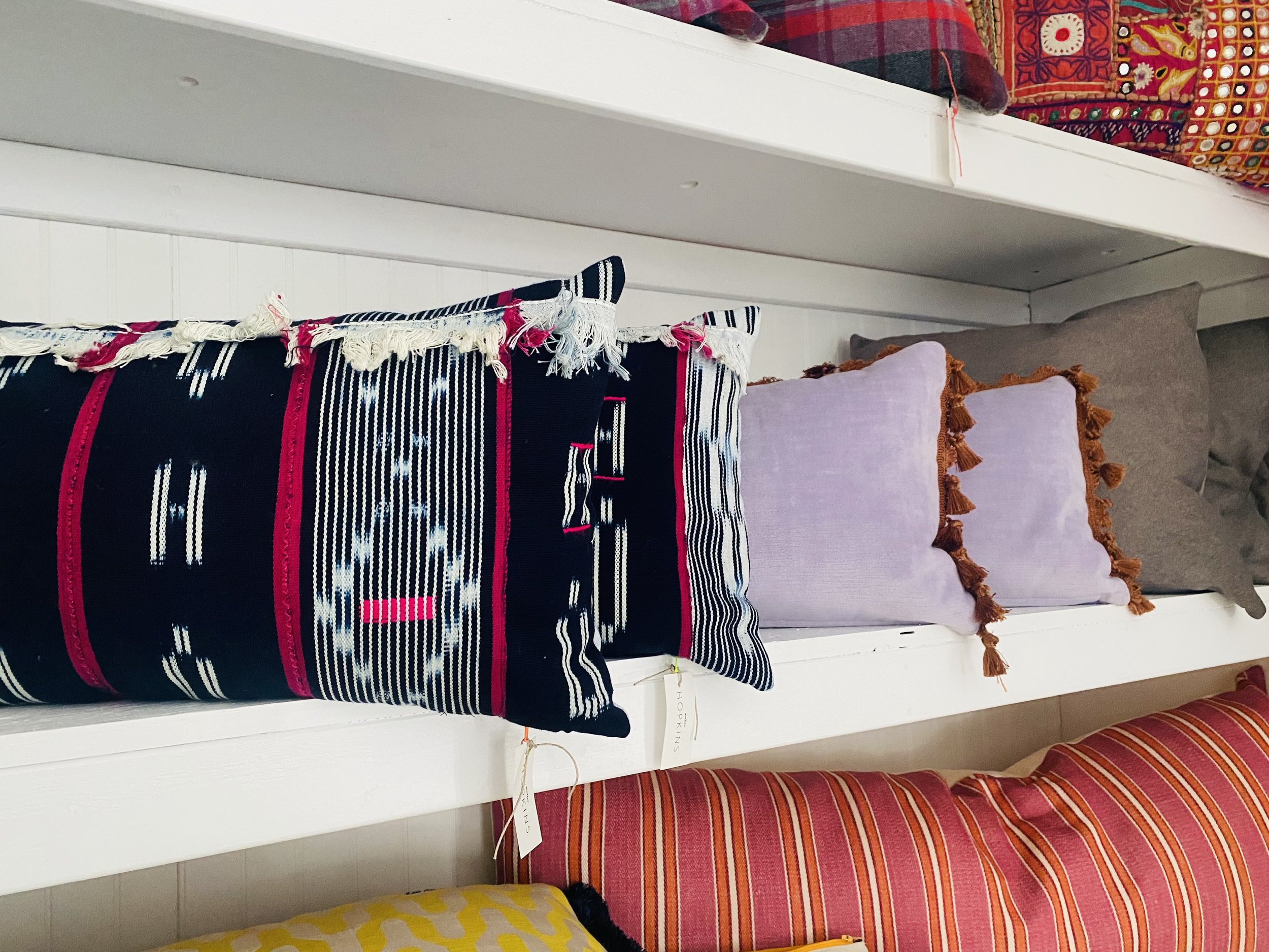 Display of decorative pillows on a white bench, featuring a black pillow with red, white, and blue patterns, light purple pillow with tassels, and brown pillow with a plain design.