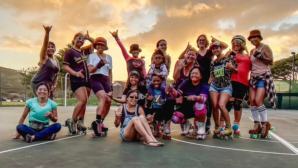 Group of women roller skating on outdoor court during sunset, some making hand gestures and smiling.