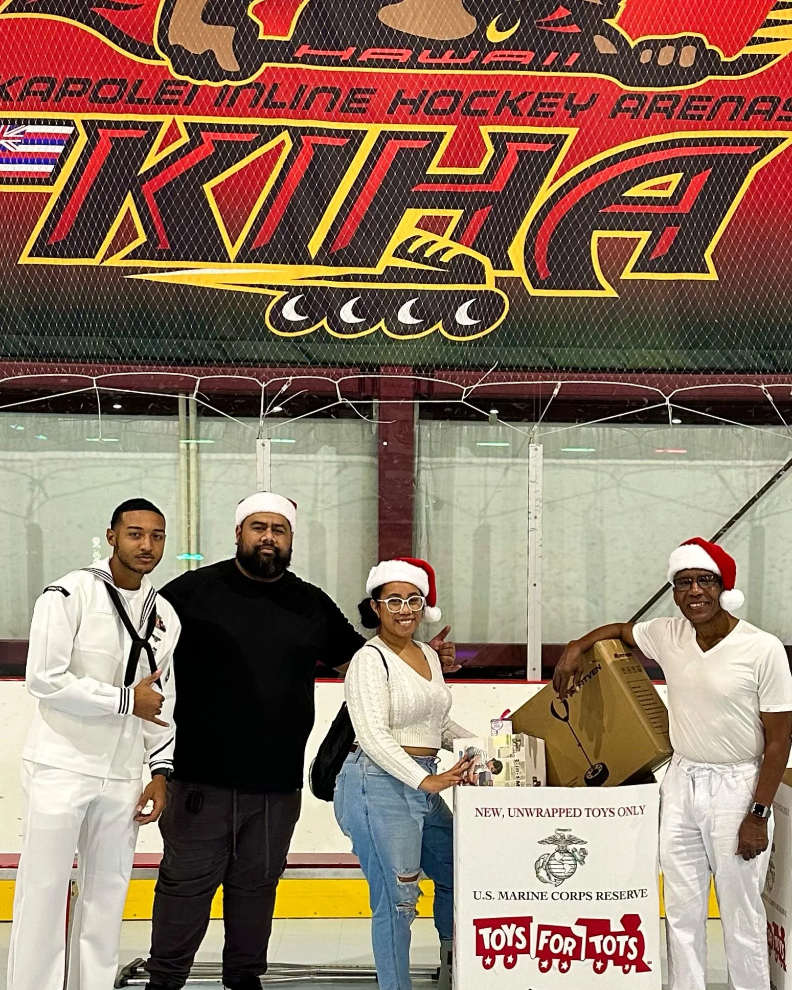 Four people wearing Santa hats and casual attire standing on an ice rink with a large Toys for Tots donation box filled with unwrapped toys, and a red and yellow hockey arena sign above them.