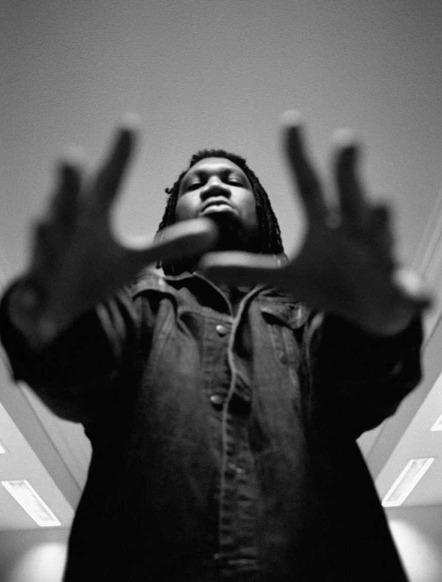 Black and white photo of a KRS-One looking down at the camera, holding their hands out in front, with an indoor ceiling visible.