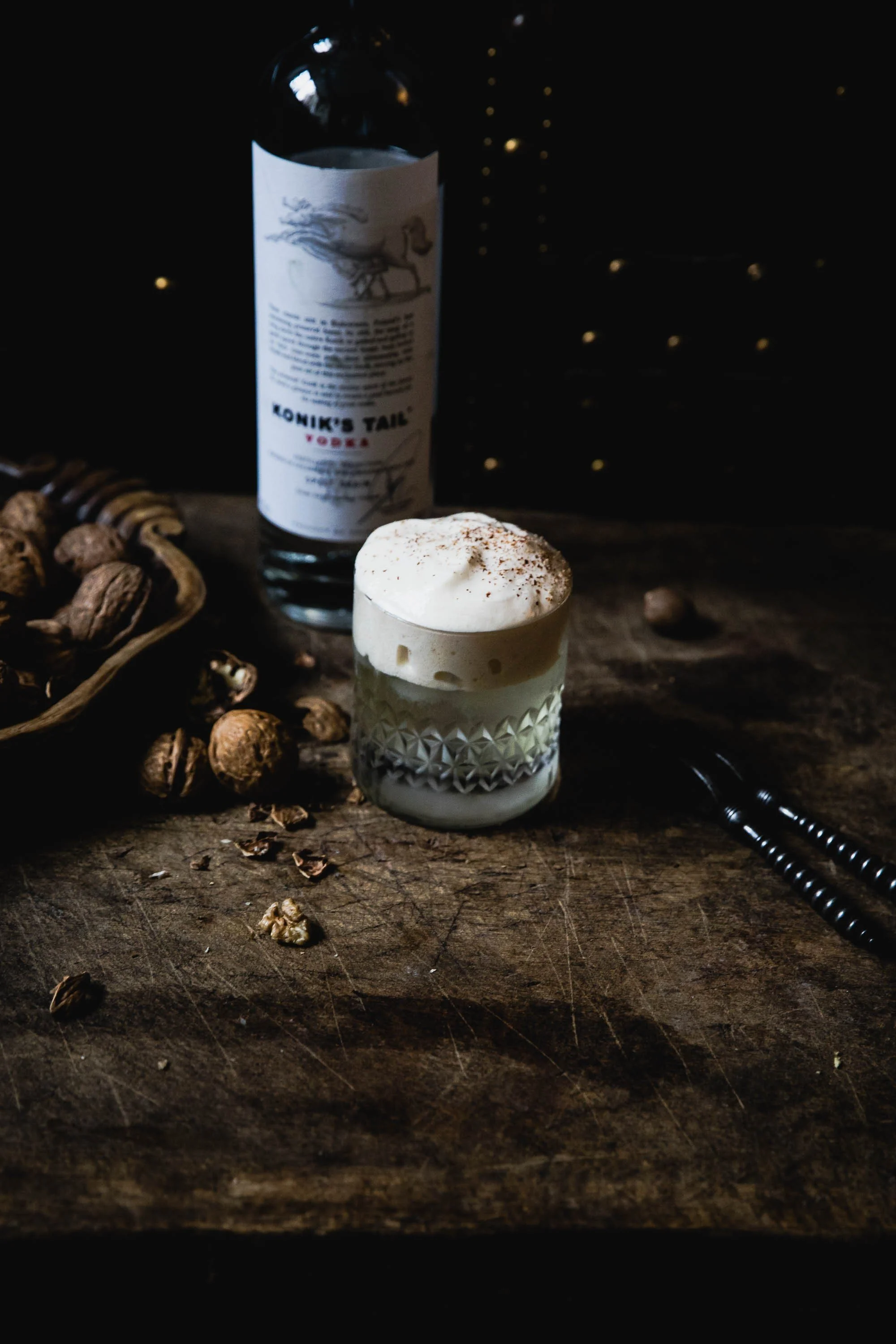A glass of whiskey with a large ice cube and an orange peel garnish on a wooden surface near a window with curtains.