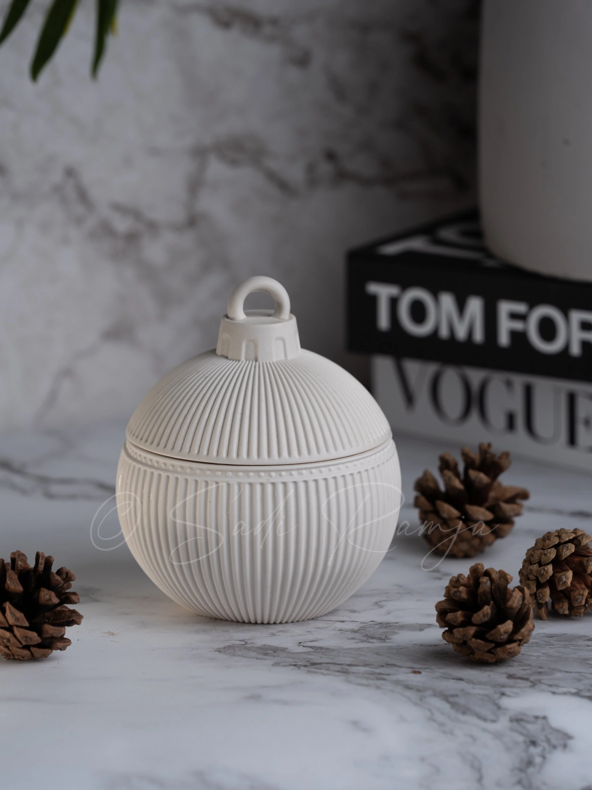 White decorative ornament with a ribbed spherical shape and a loop on top, surrounded by pine cones on a marble surface. In the background, a stack of black and white Vogue and Tom Ford books with a large white pot on top.