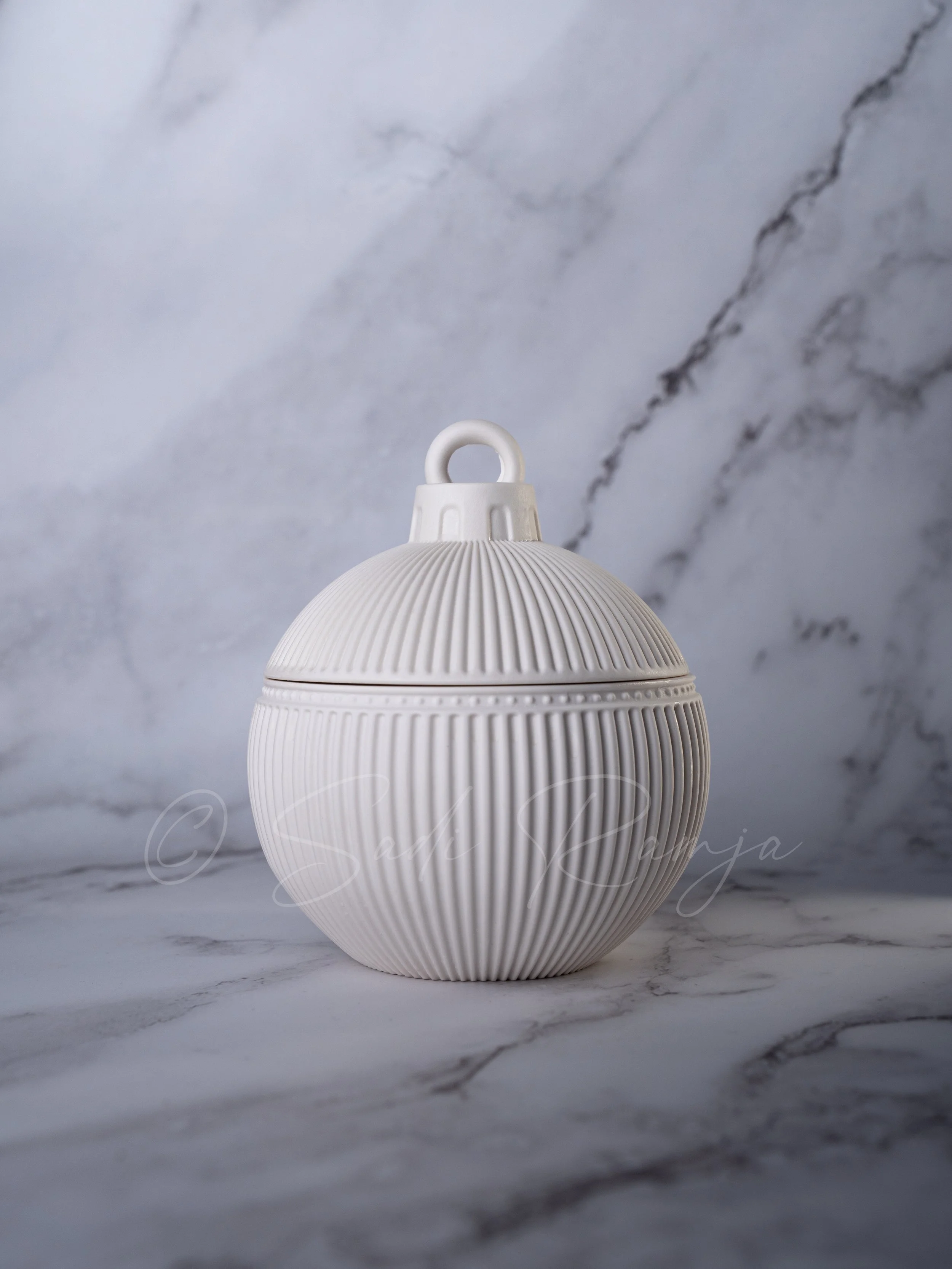 A white, round, textured ceramic container with a handle on top, set on a marble surface with a marble background.