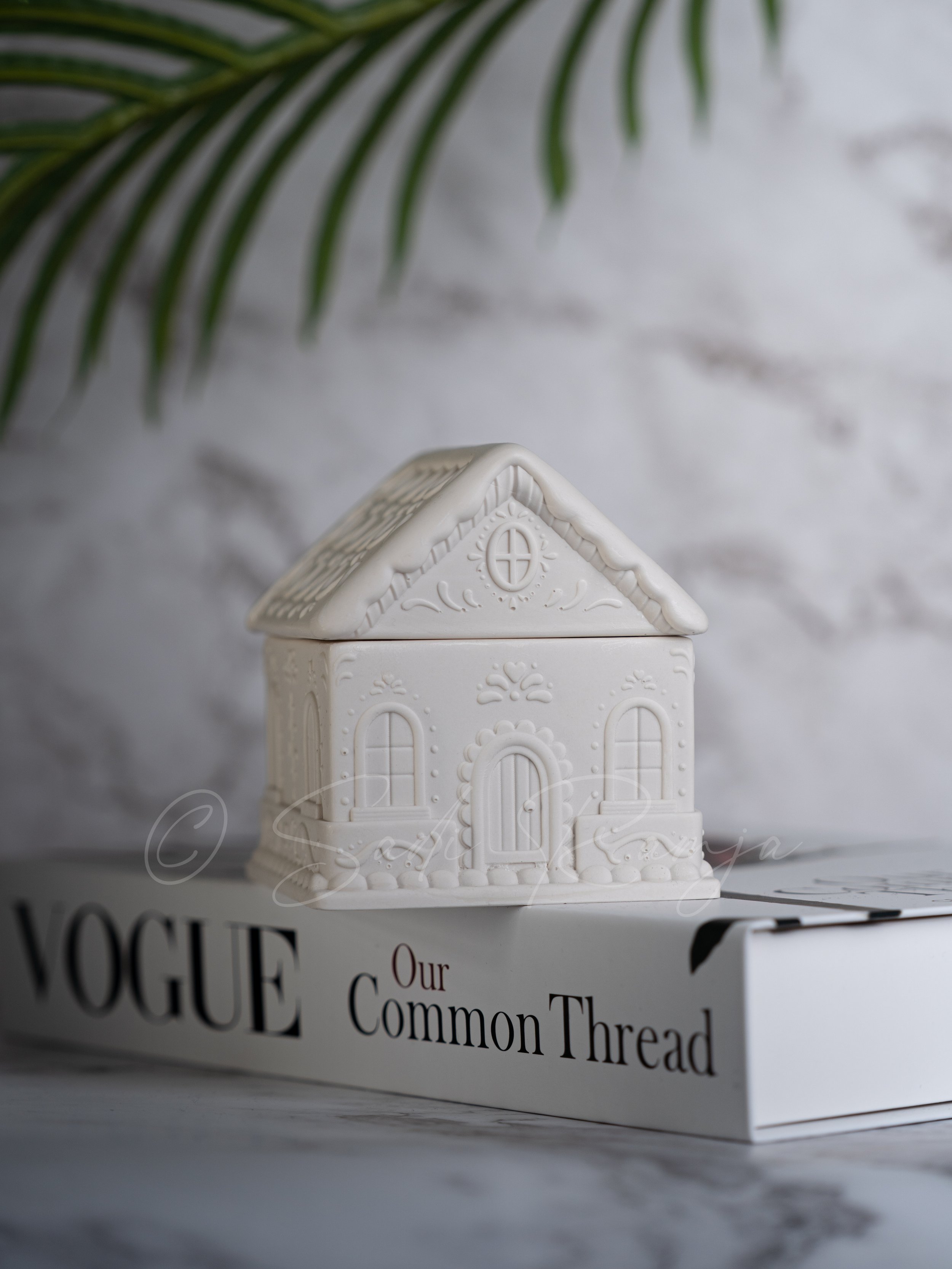 White ceramic house figurine with window and door details, placed on a Vogue magazine titled 'Our Common Thread', with a blurred outlet wall and green leaves in the background.