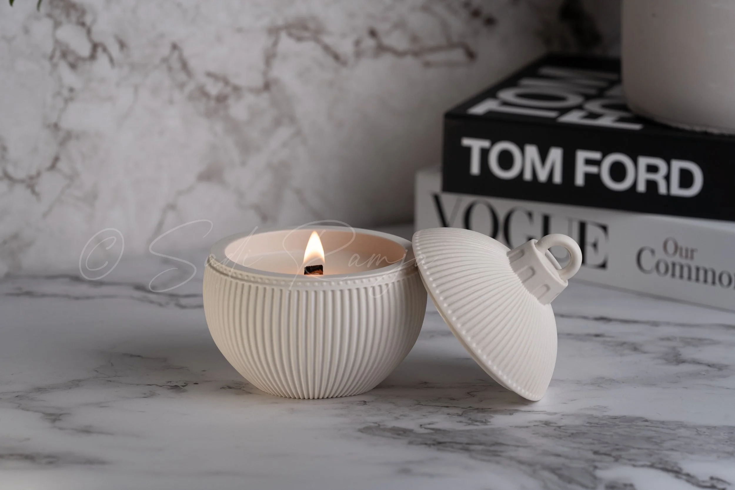 White ribbed ceramic container with a lid, containing a lit candle, on a marble surface with books in the background.