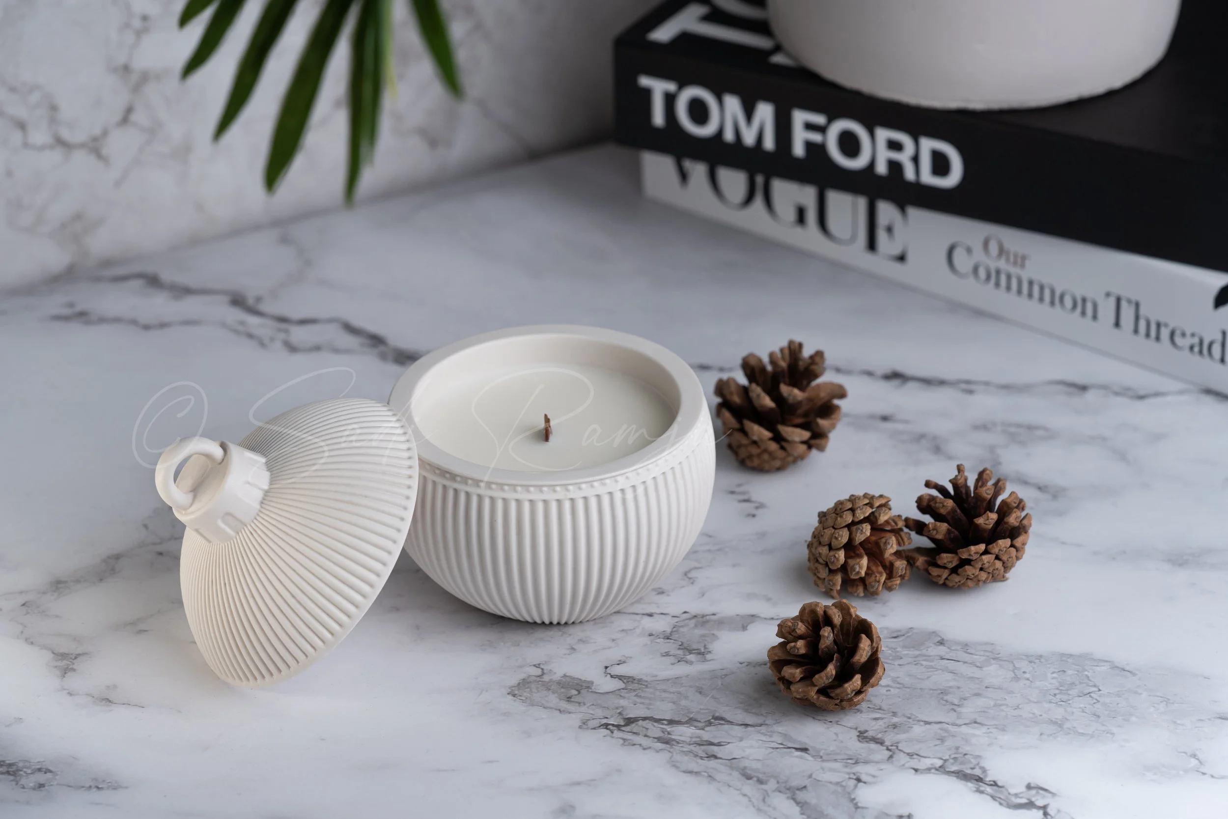 A white ceramic candle holder with a lid, containing a lit candle, placed on a marble surface with pinecones nearby, and a stack of fashion books in the background.