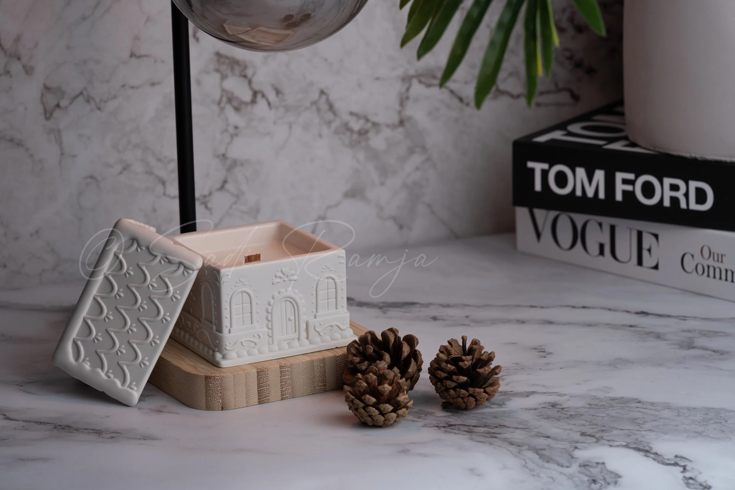Decorative ceramic house-shaped container on a wooden tray, pinecones, a black and white book titled 'TOM FORD VOGUE Our Comm', a glass sphere, and a potted plant on a marble surface.