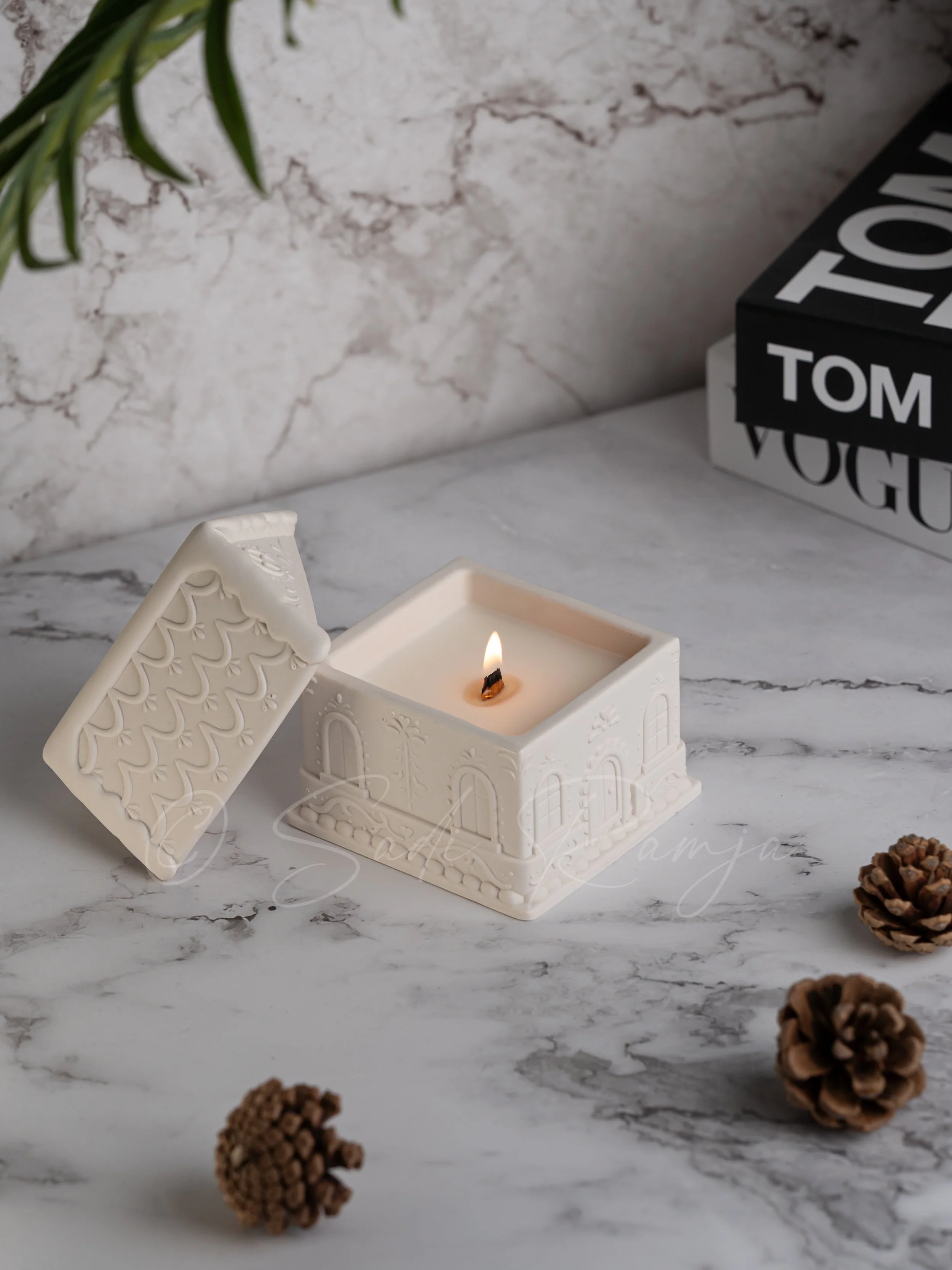 A white candle holder with a lit candle inside, designed like a house with decorative windows and doors. The lid with embossed patterns is placed nearby. Several pinecones are scattered around on a marble surface, with a gray marbled wall and a stack