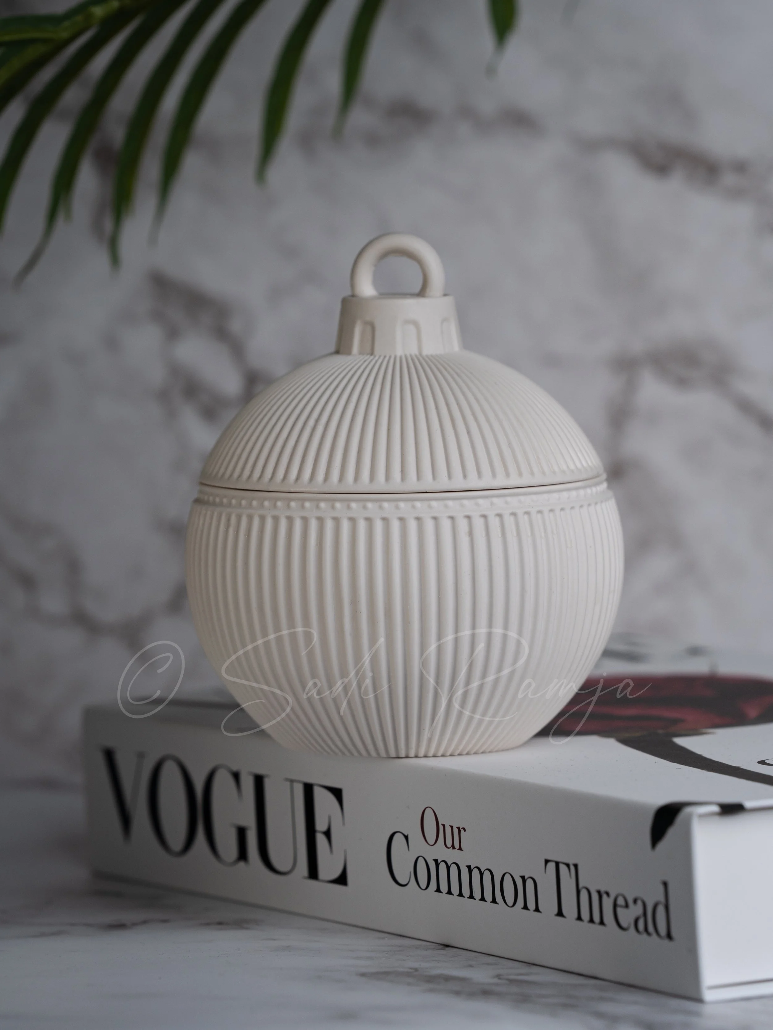 A round, ribbed, white ceramic container with a handle on top, placed on a copy of Vogue magazine with the title "Our Common Thread," against a marble and leafy background.