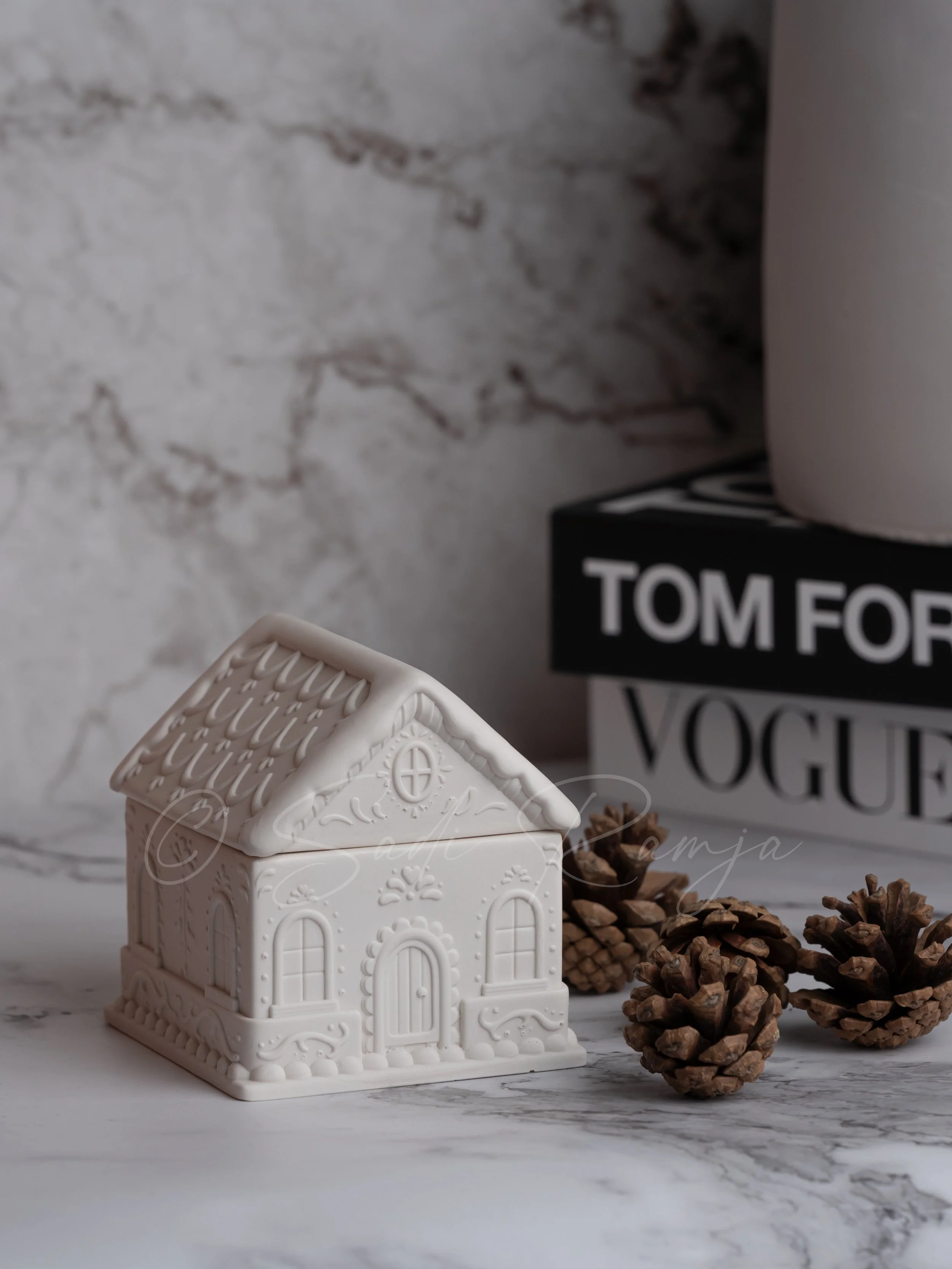White ceramic house ornament with pinecones and books in the background on a marble surface.