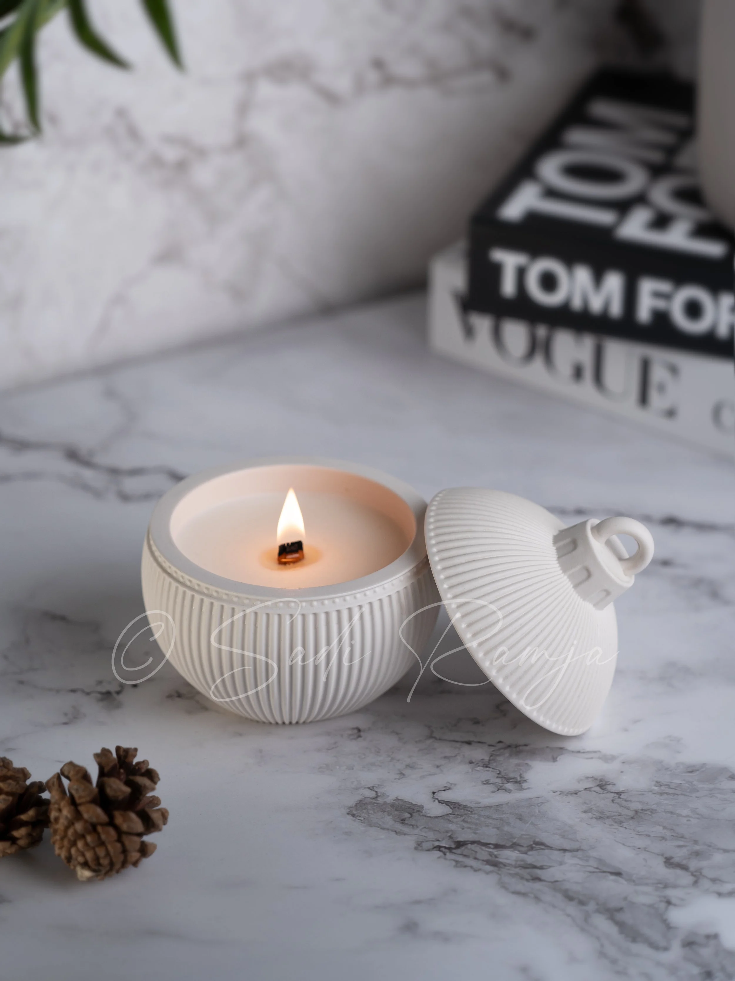 White textured ceramic candle holder with a lit candle inside, placed on a marble surface. There are two pinecones nearby, and books stacked in the background with titles including "TOM FORD" and "TOUGH."