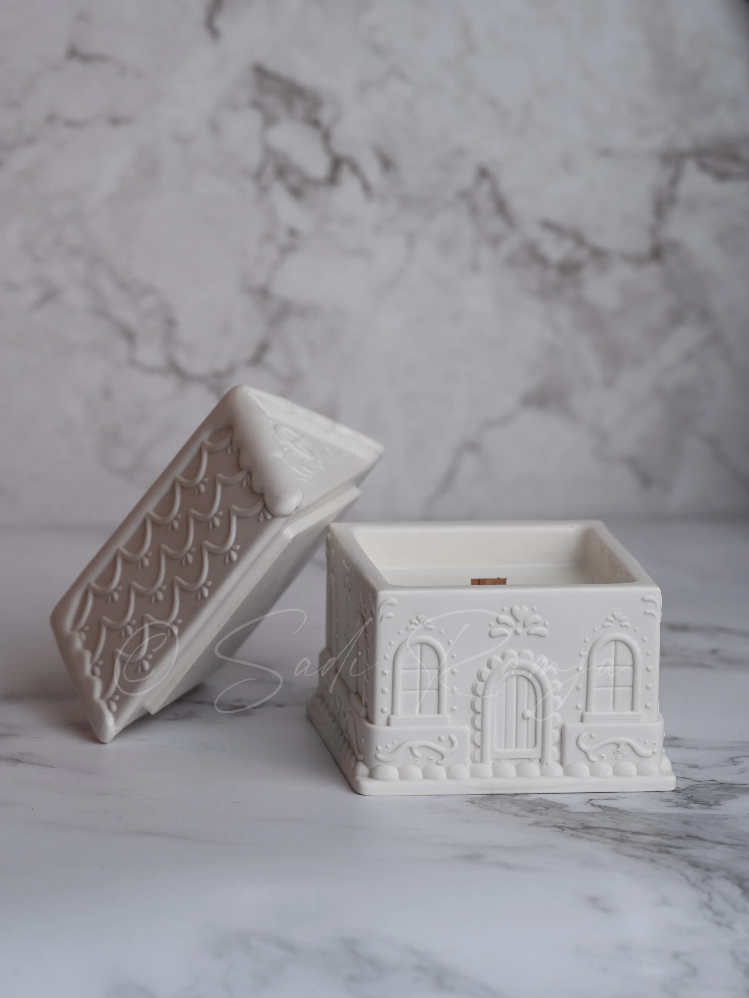 White decorative ceramic box with a roof, designed to look like a house, placed on a marble surface with a marble wall in the background.