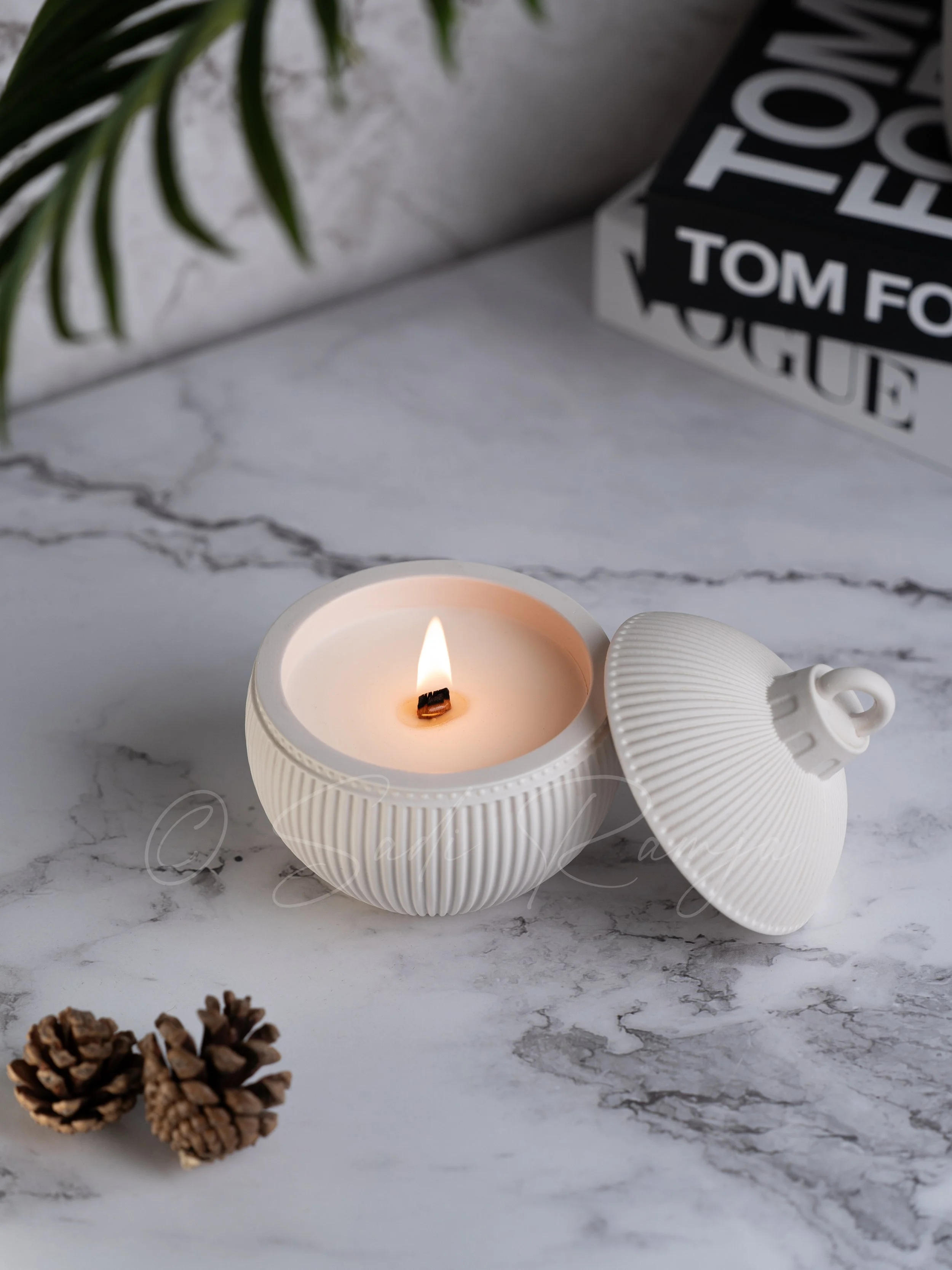 White textured ceramic candle holder with a lid, burning candle inside, on a marble surface with pinecones, a book titled 'TOM FORD' and some green leaves partially visible in background.