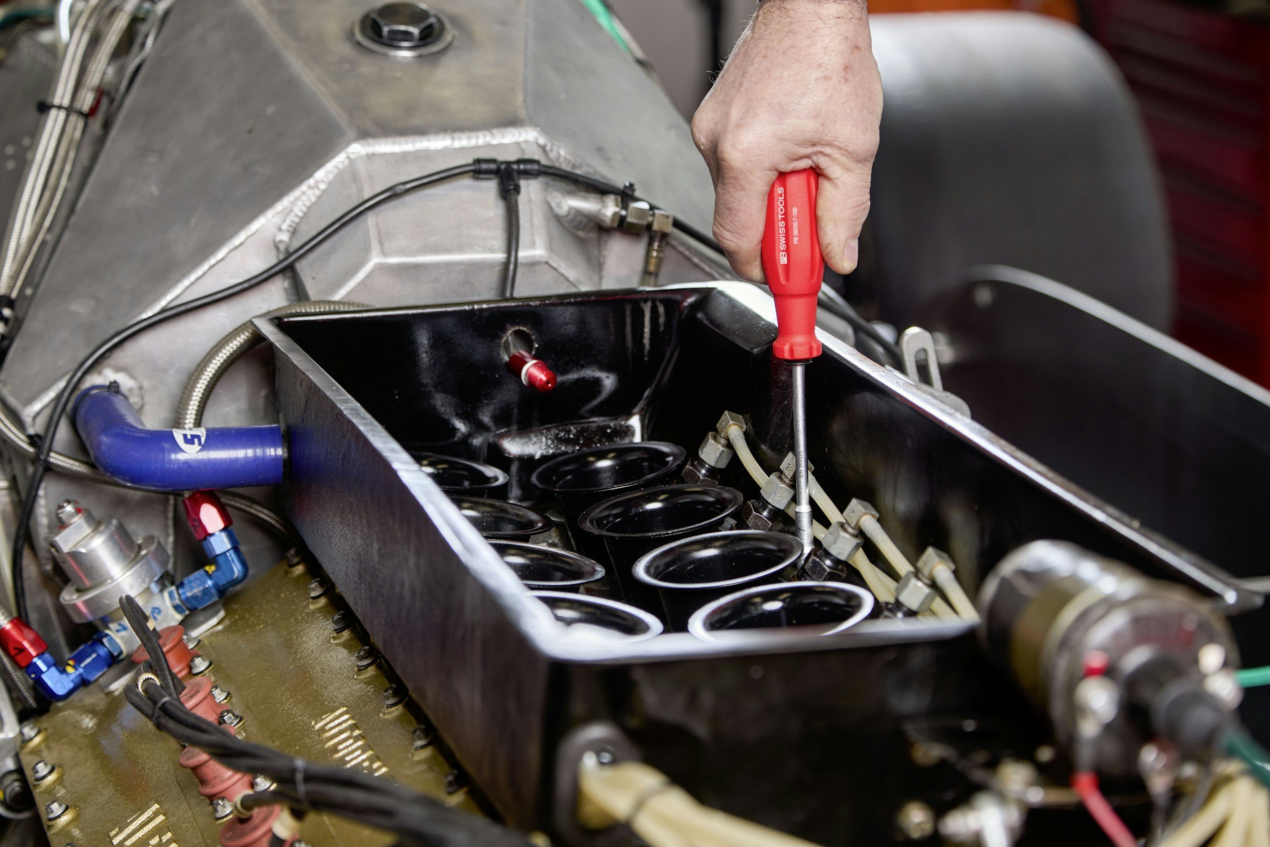 A person is using a red screwdriver to work on an engine or mechanical device, which includes multiple black circular components, wires, and metallic parts.