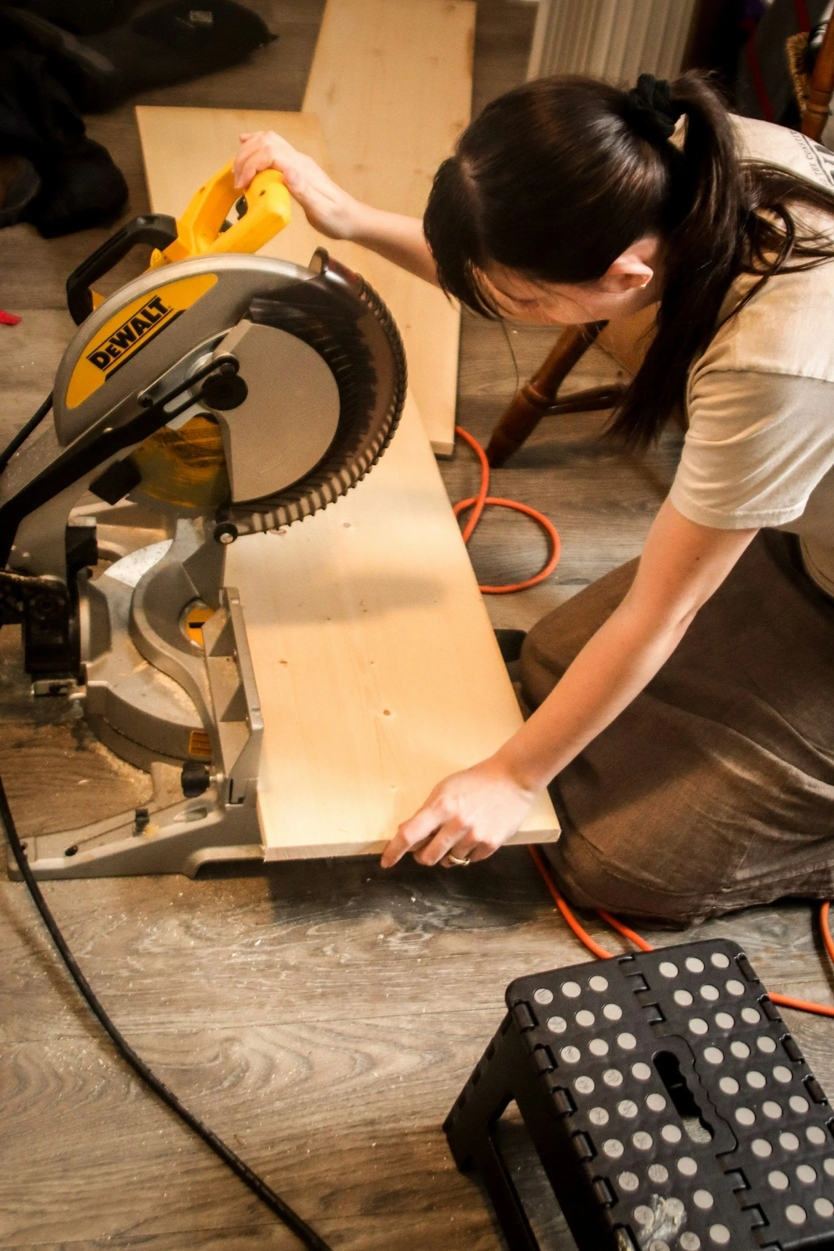 A person kneeling on the floor using a yellow DeWalt circular saw to cut a piece of wood on a table. The person is leaning forward and holding the saw with one hand. There is an orange extension cord on the floor, a black step stool with white holes, and various furniture and tools in the background.