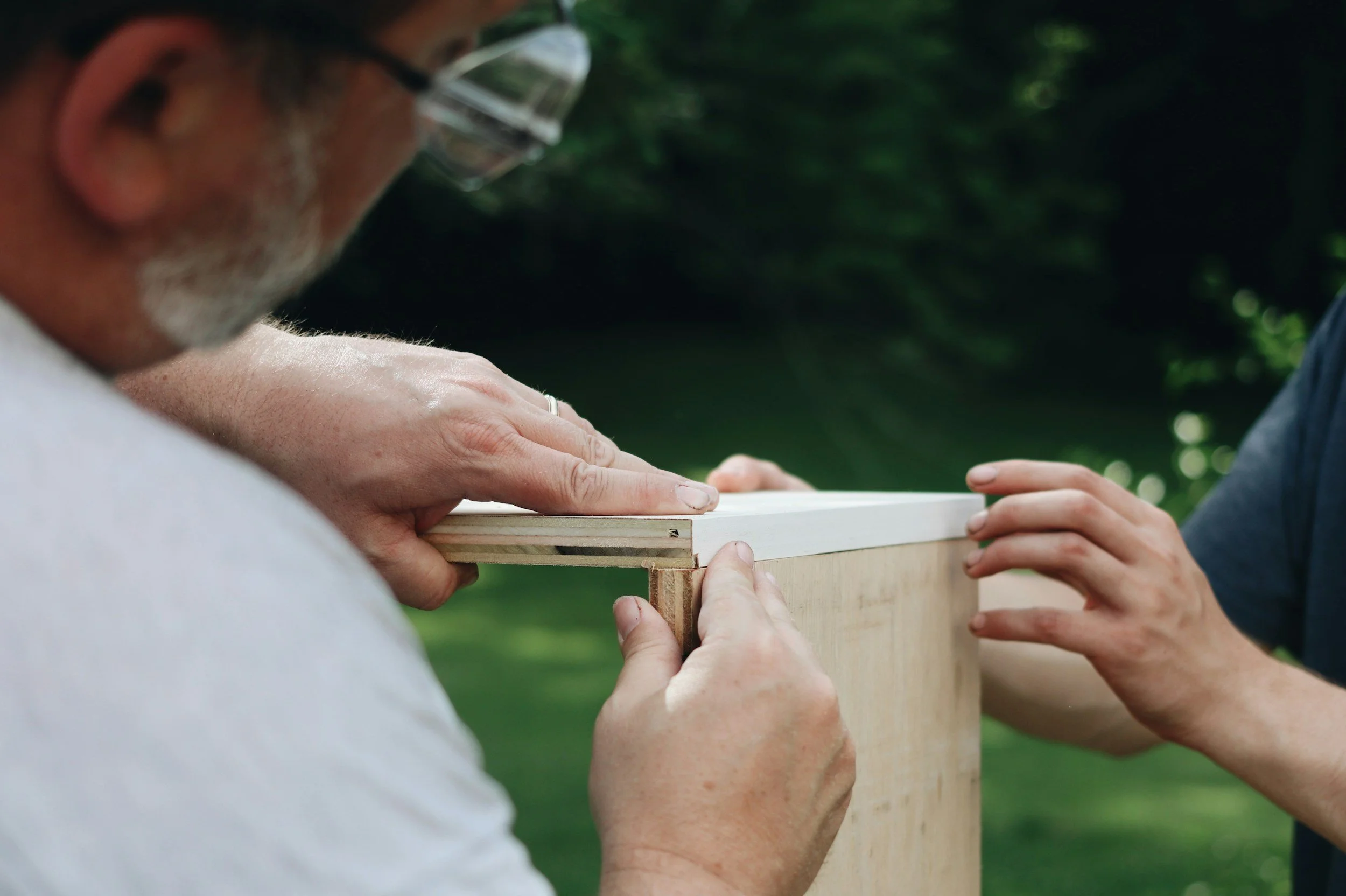 Two people working together on a woodworking project outdoors, measuring and holding a piece of wood.