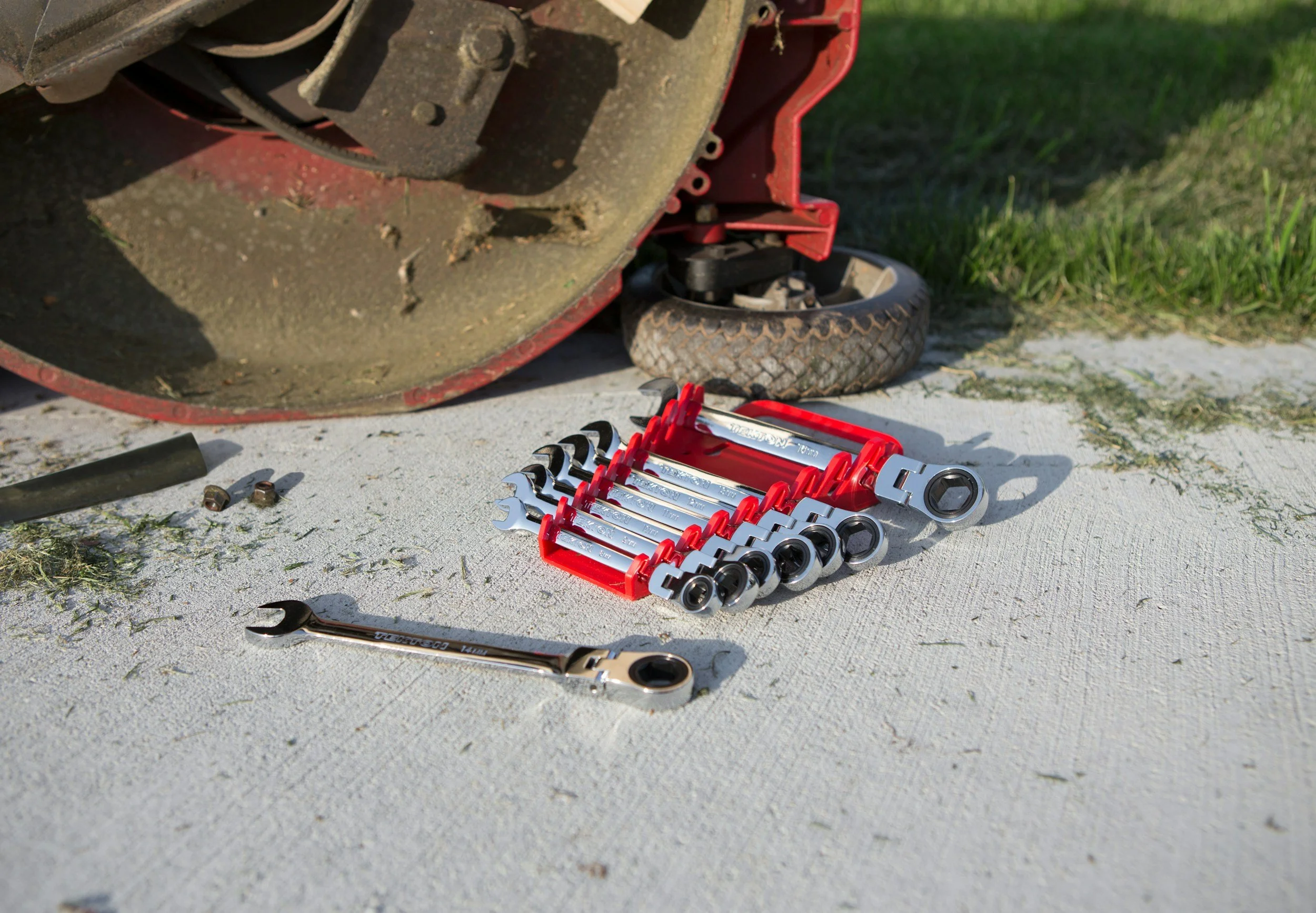 A set of wrenches arranged in a red plastic holder on a white surface outside, with a large, rusty power tool and some scattered nuts nearby.