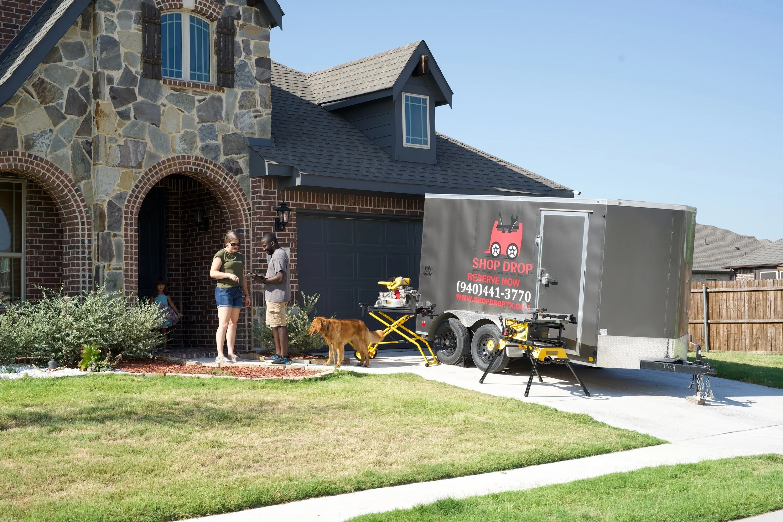 Residential house with stone and brick exterior, two visible windows, and a garage. In the driveway, three people and a dog are present, with a mobile workshop or service trailer labeled "Shop Drop" parked nearby. The scene is set in a suburban neighborhood on a sunny day.
