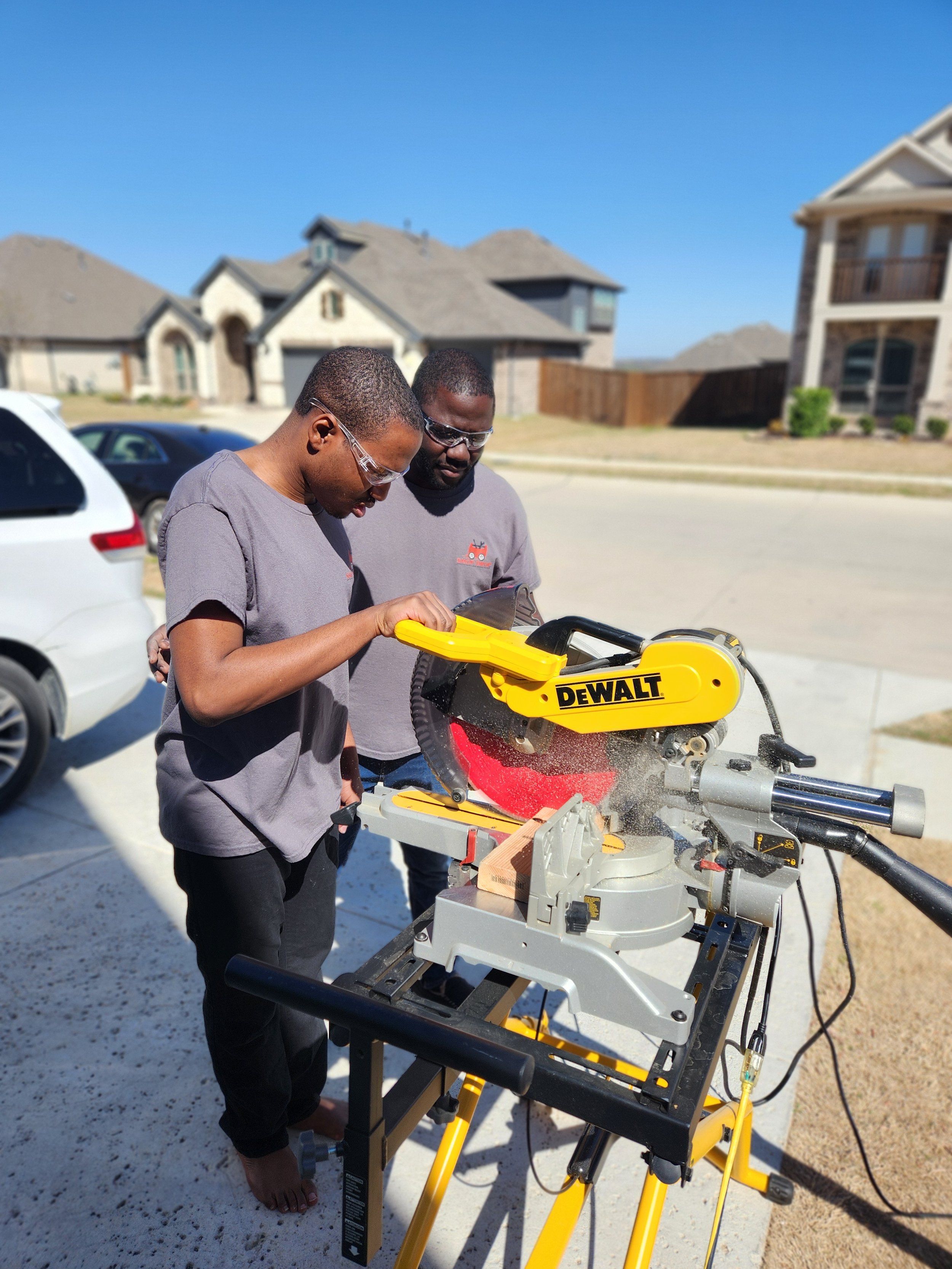 Two men operating a yellow DeWALT miter saw outside in a residential neighborhood with houses in the background.