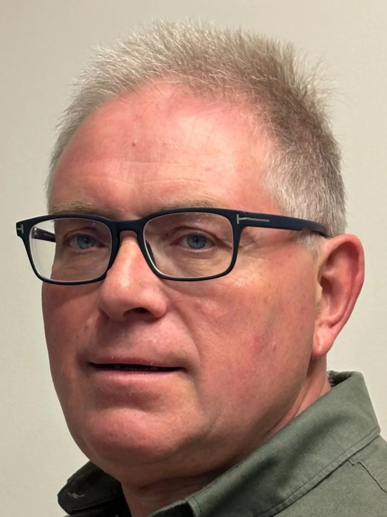 Smiling elderly man wearing glasses and a black jacket indoors, with two other people in the background.