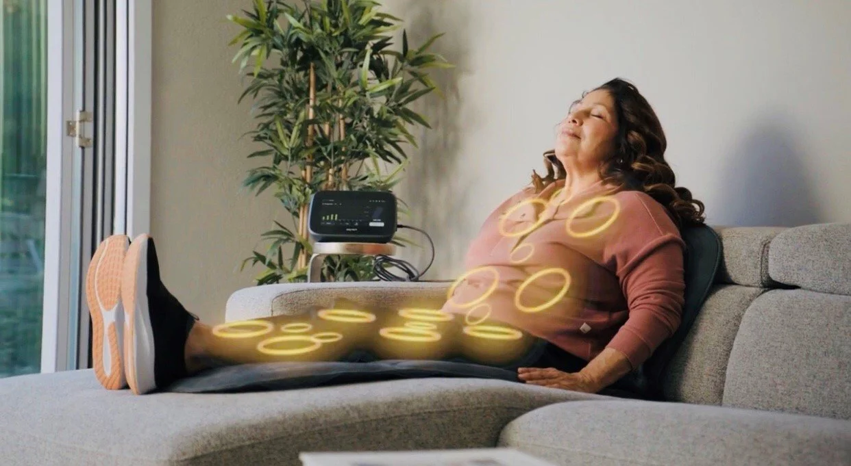 Woman sitting on her couch with her legs up while getting a  BEMER Therapy session while laying on the pad. The rings  overlaying her body depicts the treatment areas.