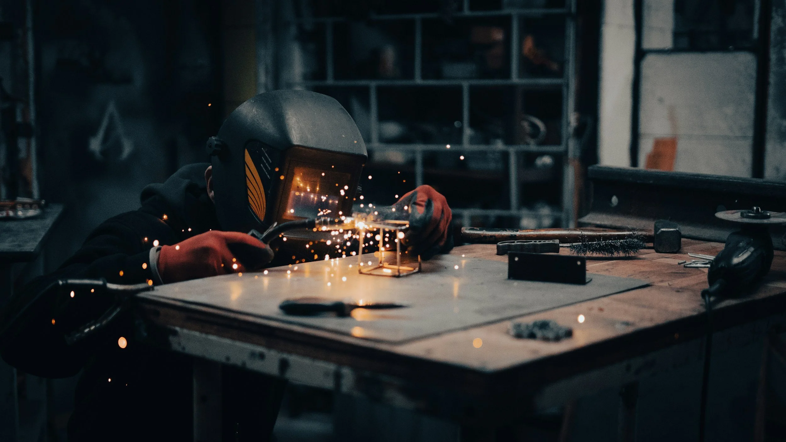 A person welding metal in a workshop, wearing a welding helmet and gloves, with sparks flying from the welding process.