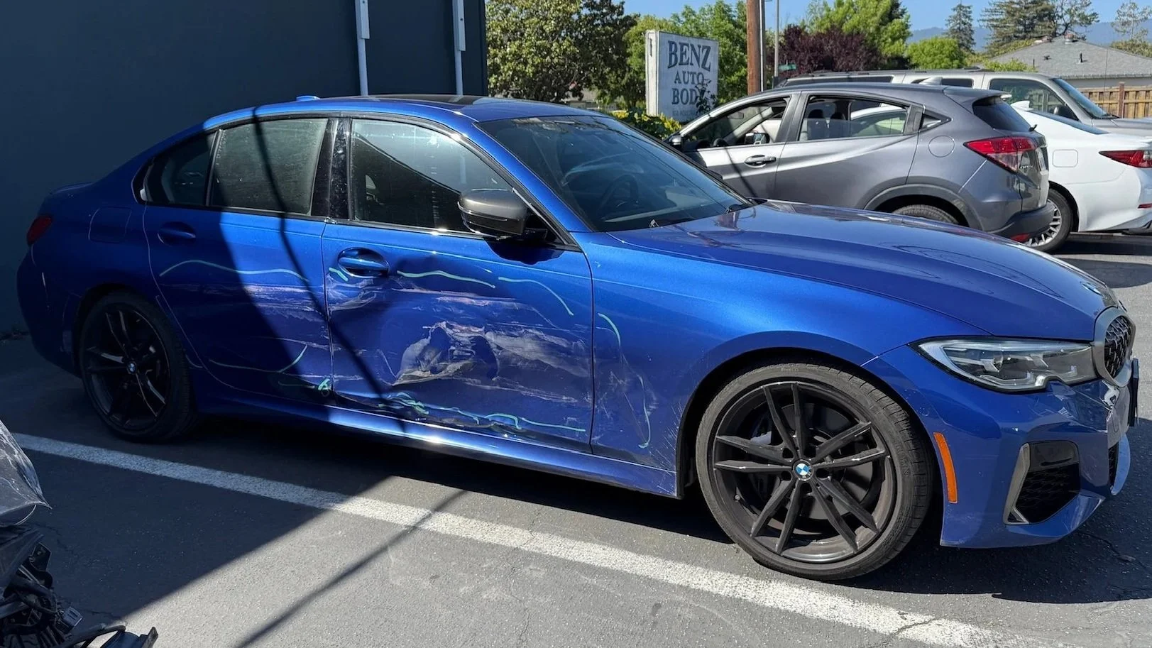 Blue BMW sedan with noticeable damage on the driver's side door and rear door, parked in a lot with other vehicles. The scene is outdoors on a sunny day.