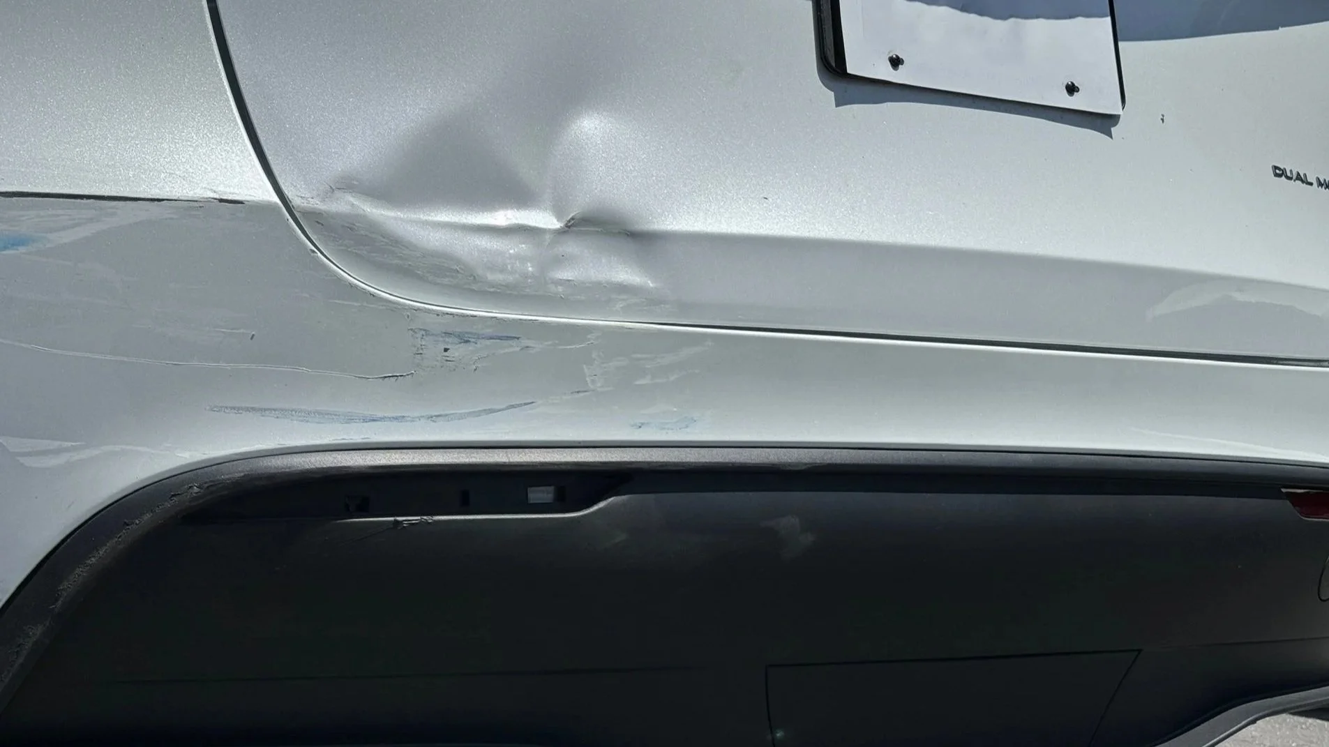 Close-up of a silver car with visible damage on the front left side, including a large dent and scratches, with part of a license plate visible at the top.